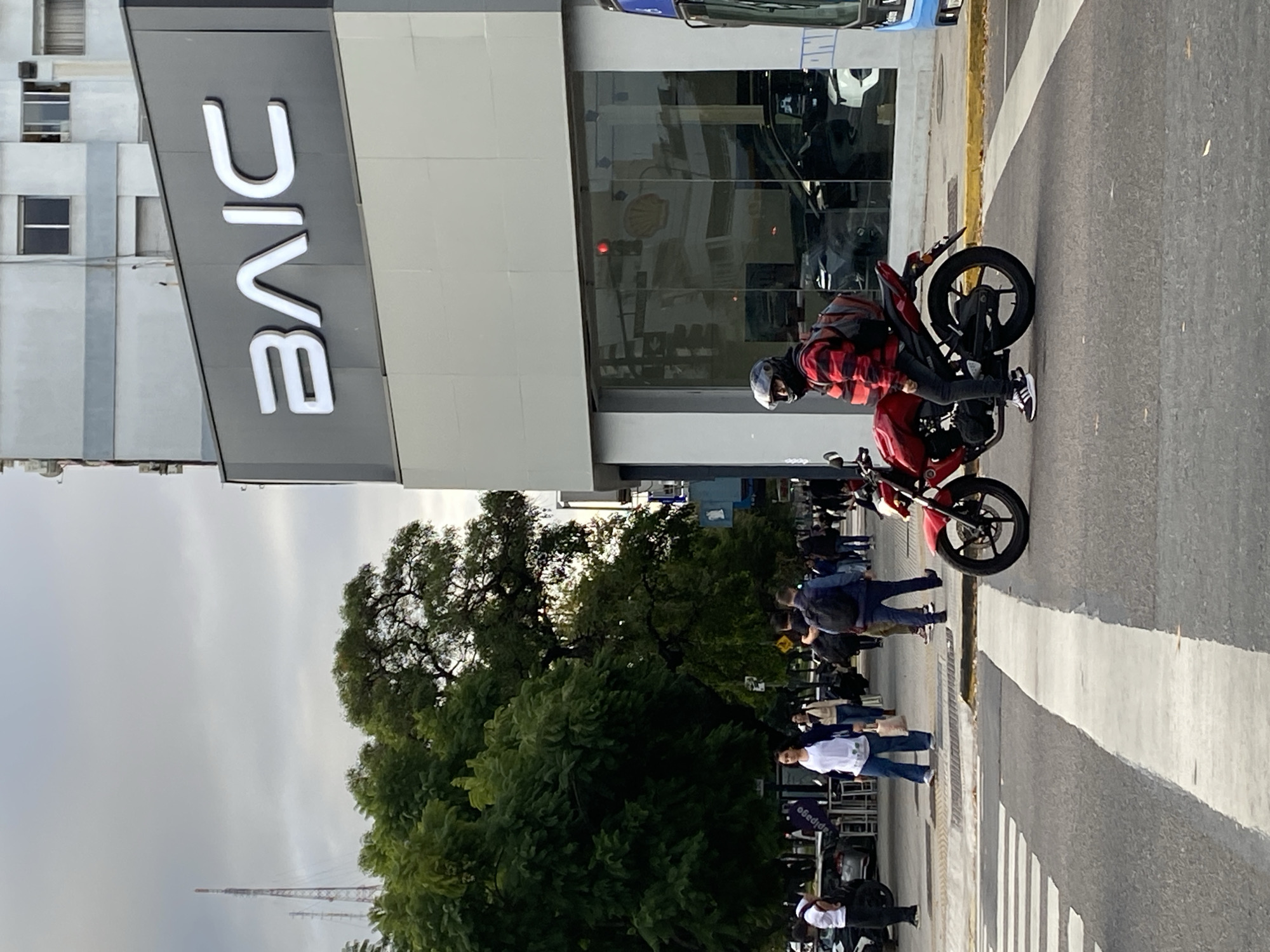 The image depicts a scene in Buenos Aires, Argentina. A motorcyclist wearing a helmet and a red plaid shirt is stopped on a red motorcycle on a street with crosswalks. People are walking on the sidewalk and crossing the street near the motorcycle. In the background, a building with a large "BAIC" sign is visible. A radio transmission tower pierces the sky in the distance.