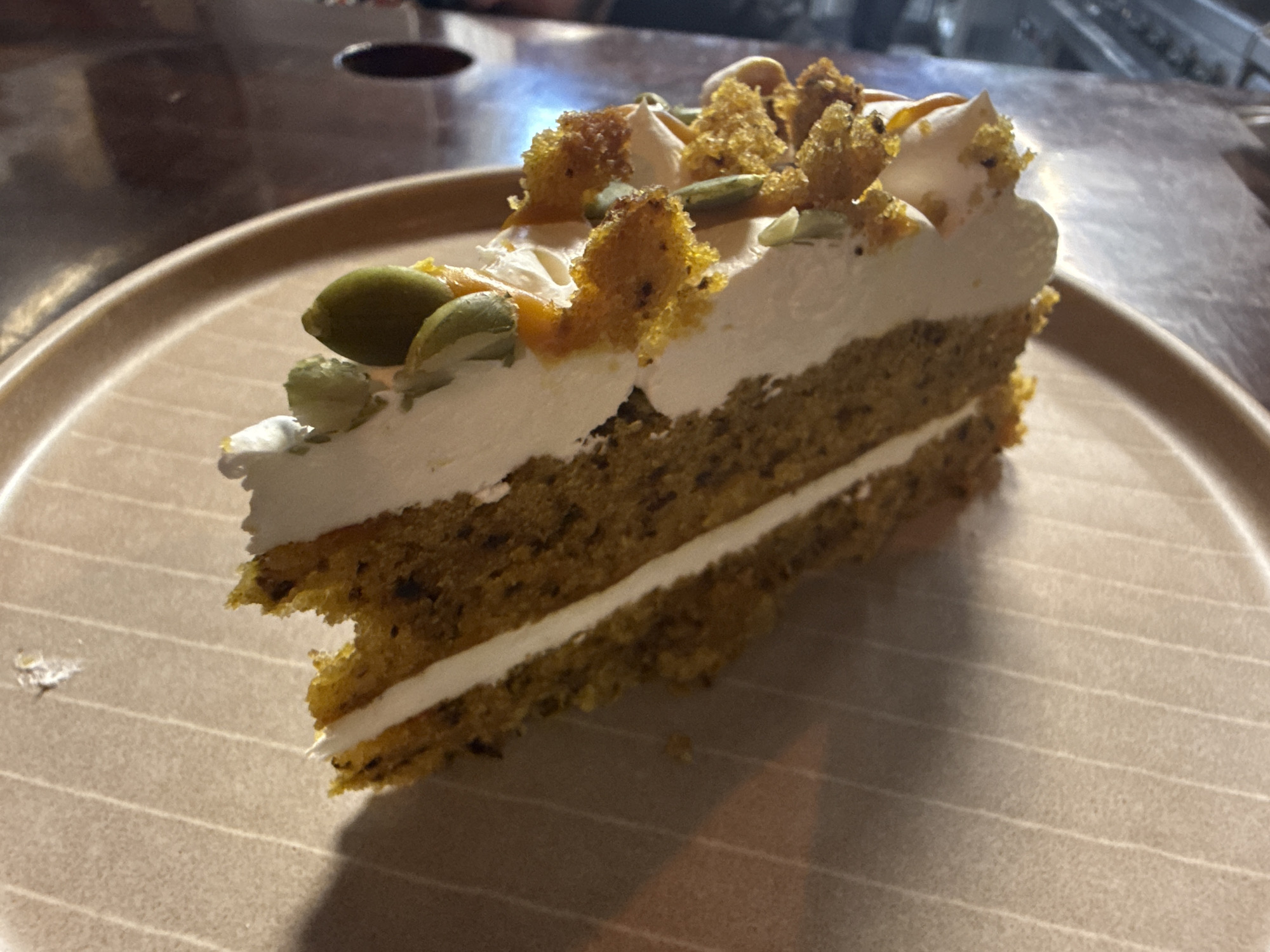 A close-up shot depicts a slice of cake on a beige plate with subtle, parallel lines. The cake features two layers of dark, speckled cake separated by a layer of white frosting. The top of the cake is adorned with a thick layer of white frosting, topped with crumbled cake pieces and pumpkin seeds. The environment is dimly lit. The image does not provide any information about the location, city, or country.