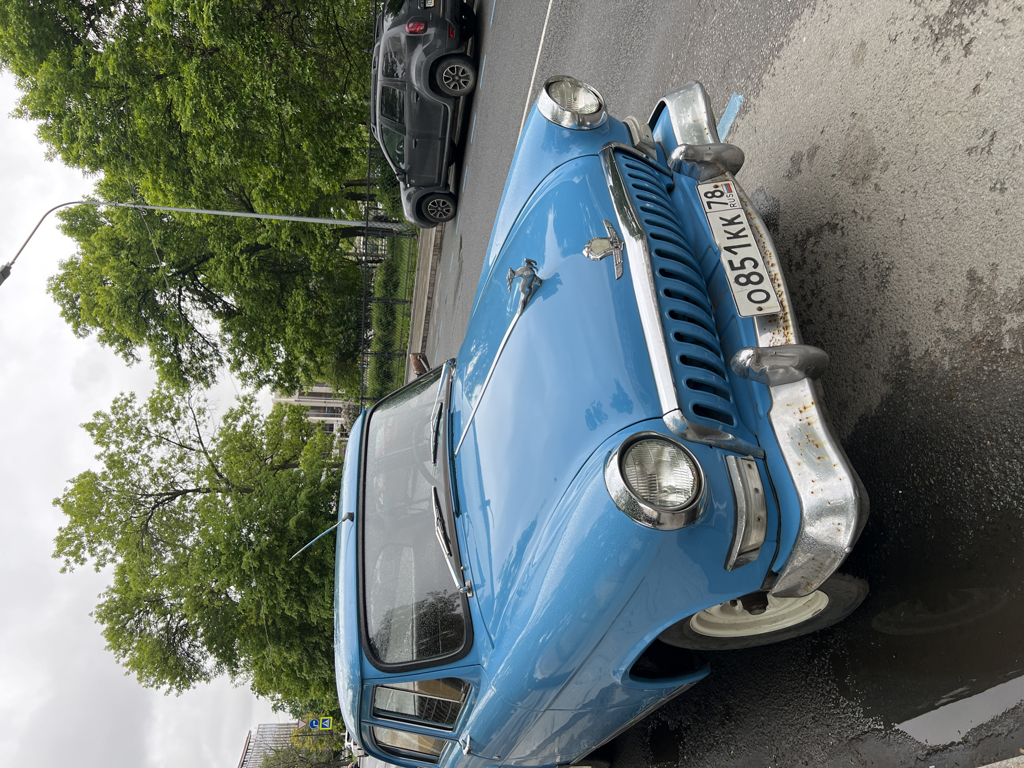 A close-up, slightly rotated, eye-level shot depicts a light blue GAZ-21 Volga, an automobile produced in the Soviet Union from 1956 to 1970, parked on an asphalt road in Saint Petersburg, Russia. The license plate "O851KK 78 RUS" is prominently displayed on the front of the car. Visible details include chrome bumpers, headlamps, a chrome hood ornament, and windshield wipers. The car is wet, suggesting recent rain. In the background, partially obscured by the car, is a dark SUV. Trees and a street light are visible in the background. The lighting indicates a cloudy, overcast day. The overall environment suggests an urban setting with a mix of parked cars and street vegetation.