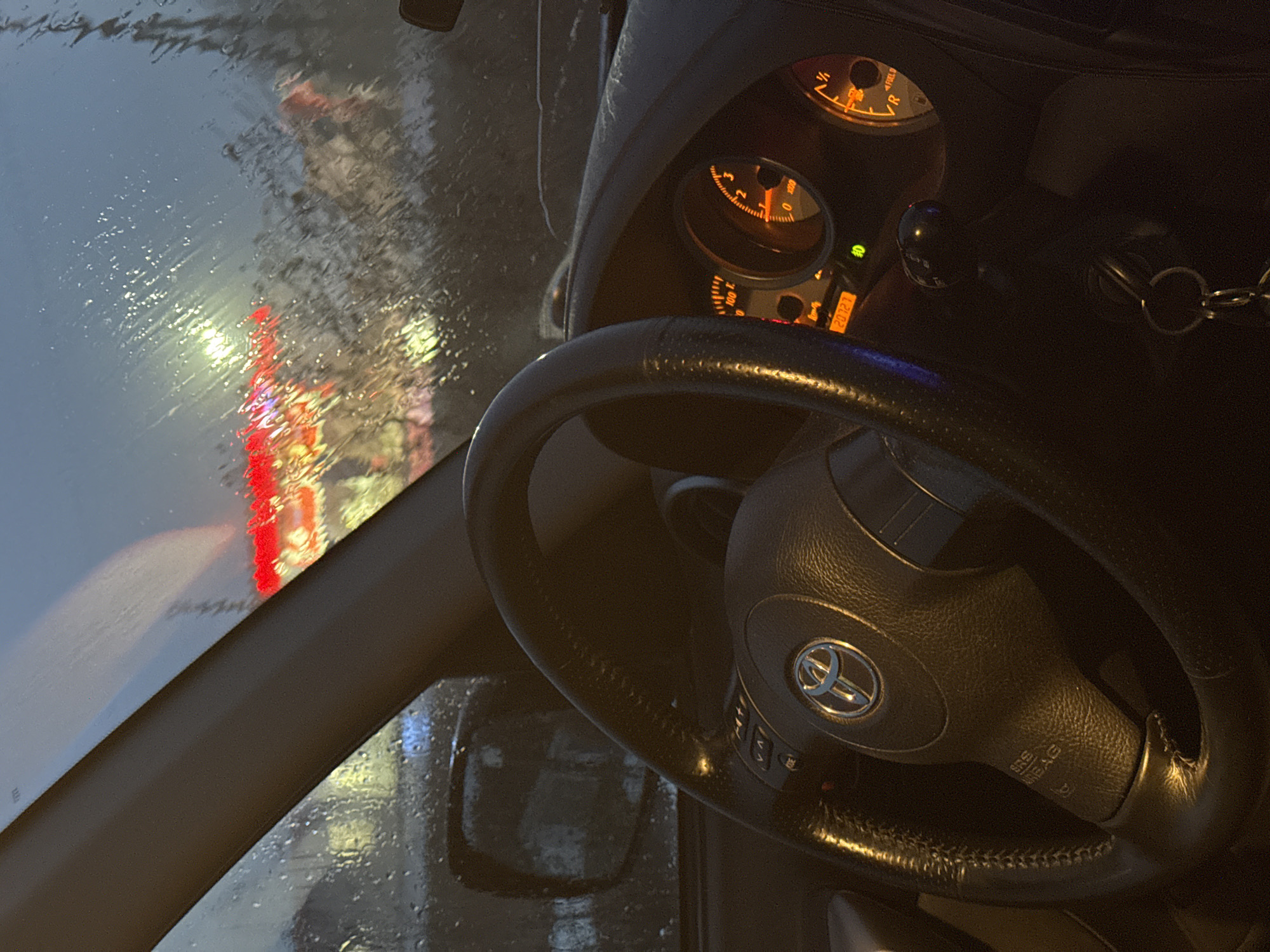 The image shows the interior of a car, specifically the driver's seat perspective. The steering wheel is prominently featured, with a Toyota logo visible at the center. The dashboard gauges are illuminated, including the speedometer and fuel gauge. Outside, the windshield is covered with raindrops, obscuring the view. Red and white lights are visible through the windshield, suggesting city lights or traffic signals. The side view mirror is visible but the scene outside is not clear. No people are visible. The location of Yazlova, Russia is not identifiable from the image.
