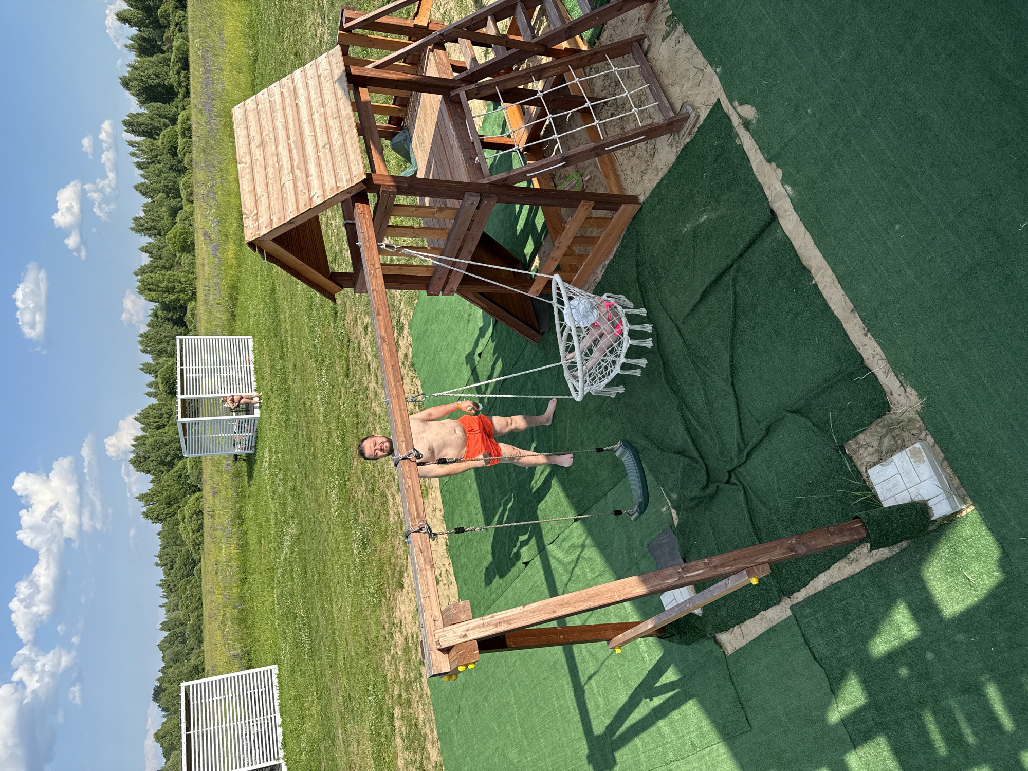 In the city of Koloksha, Russia, on a bright, sunny day, a man with a beard, wearing orange swim trunks, stands barefoot on a green artificial turf area. He is positioned near a wooden swing set, part of a larger wooden playground structure. The playground features a slide, a climbing wall with rope netting, and a wooden fort with a roof. A white macrame swing with a child inside is suspended from the swing set. To the left of the playground, another white structure with horizontal slats is visible, and further in the background, a second similar structure houses a person. The surrounding environment is a grassy field with scattered trees and a line of trees in the distance under a blue sky with white clouds. The ground beneath the playground is sand, and the artificial turf is laid out in sections.
