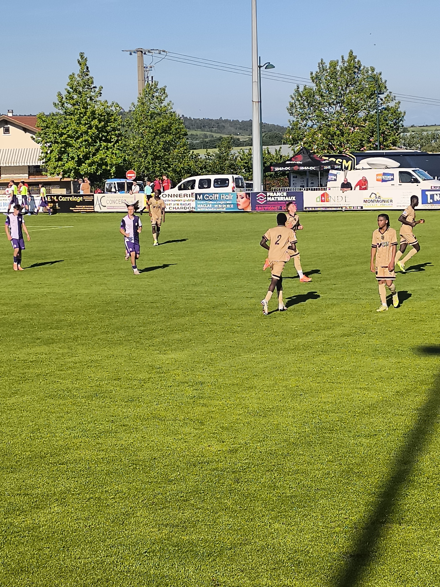 The image shows a youth soccer game in progress in Maclas, France. A group of players wearing white and purple uniforms are on the left, while players in brown and black uniforms are on the right. There are spectators visible behind the field perimeter. The scene is set on a bright, sunny day, with green grass covering the playing field. Advertisements for local businesses are visible along the perimeter of the field, including "M Coiff Hair Maclas." Buildings, trees, and hills are visible in the background.