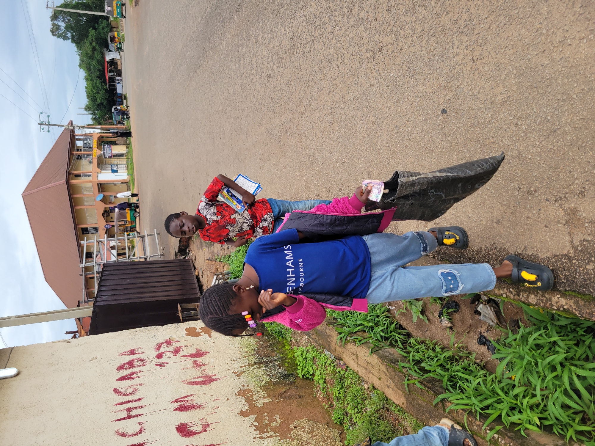 The image captures a street scene in Jos, Nigeria. Two children are walking along a dusty road. The girl in the foreground, facing away from the camera, wears a blue t-shirt with the text "GENHAMS MELBOURNE" and a pink jacket. She carries a black bag and appears to be holding some money. The boy behind her, also facing away, wears a red patterned shirt and blue jeans, and holds a book. The environment includes a wall with red markings on the left, a metal fence, lush green vegetation, and a paved road with some debris. In the background, there are buildings, utility poles, and several motorized rickshaws, commonly known as Keke Napep, typical of Nigerian streets. The sky is overcast, suggesting a cloudy day.