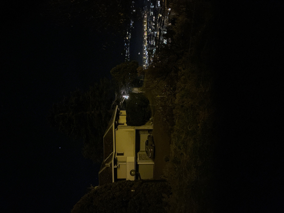 This nighttime photograph captures a serene elevated view overlooking a city in Sondrio, Italy. In the immediate foreground and midground, a multi-story building, likely residential, stands on the right side of the frame with light-colored walls and visible outdoor utility units. A vehicle is partially parked near the building. A paved path or driveway extends from the structure, flanked by dense, dark vegetation and trees that border the scene on both sides.
Beyond the immediate surroundings, a vast expanse of distant city lights shimmers horizontally across the upper portion of the image. These numerous artificial illuminations suggest a sprawling urban area nestled in a valley or across a wider plain, creating a striking contrast with the dark foreground. The night sky, a deep black, dominates the upper-left segment of the photograph, featuring a single, distinct bright star or planet.
The image conveys a quiet, tranquil atmosphere, highlighting the beauty of urban lights from a remote, elevated perspective. There are no people visible, and no specific activity is taking place. The scene emphasizes the natural darkness of the night interrupted by the glow of distant human habitation.