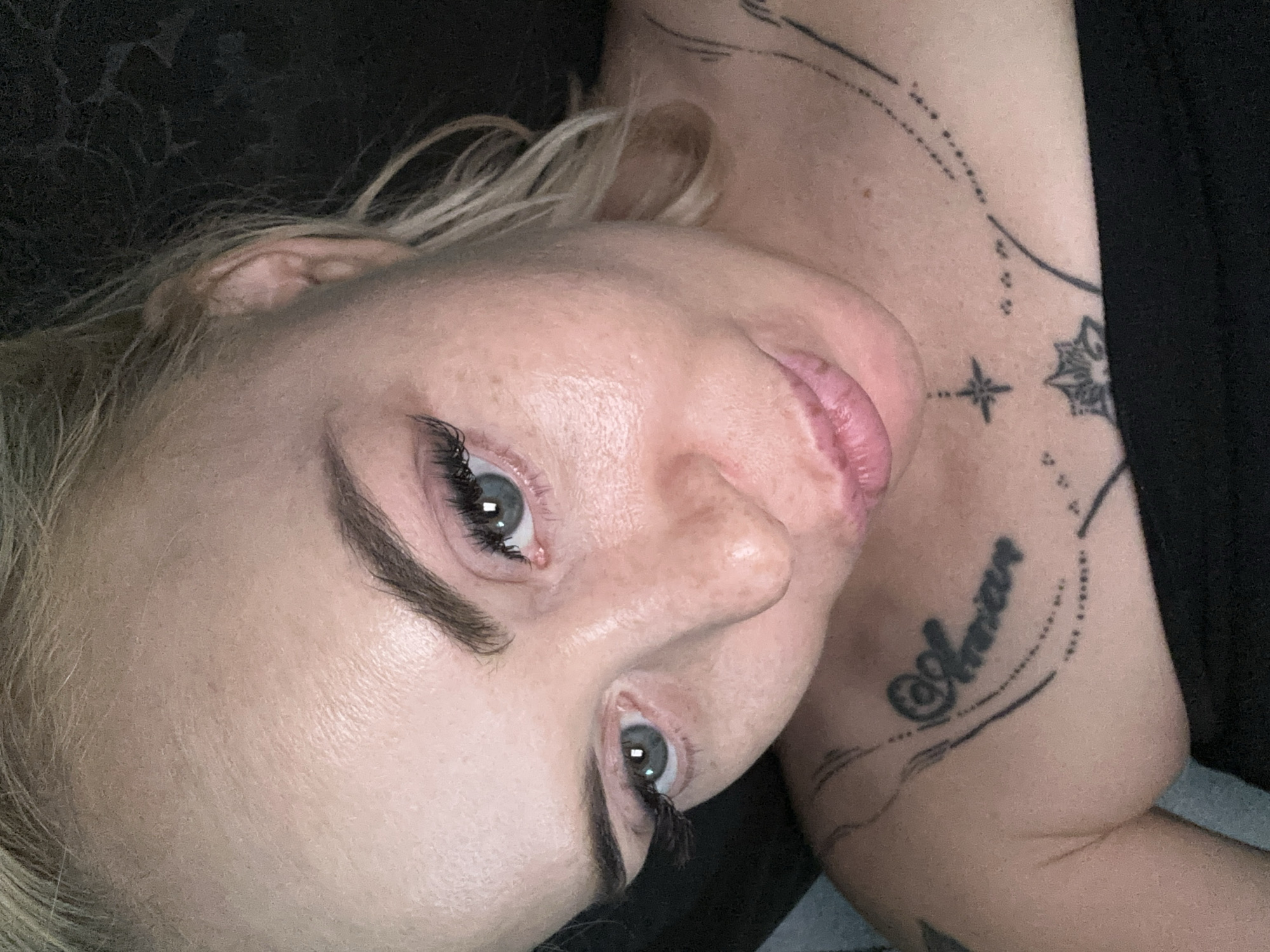 A close-up, high-angle portrait features a fair-skinned female individual with blonde hair and blue-grey irises. Her eyebrows are well-defined, and her eyelashes are prominent. The skin on her face exhibits visible texture, including fine lines on the forehead and a slight sheen on the cheeks and nose, indicative of skin oils or lighting. Her lips are full and slightly parted, forming a subtle pout.
She is wearing a simple black garment, possibly a strapless top, which covers her chest. Multiple tattoos are visible on her upper torso:
* On her left shoulder/collarbone area, a script tattoo, resembling "Amanar," is enclosed within a dotted oval design.
* Centered on her upper chest, below the collarbones, is a four-pointed starburst tattoo.
* Further down on the sternum area, a stylized floral or mandala pattern is tattooed.
* Thin, flowing lines and scattered dots connect these designs and extend across her upper chest and shoulders.
The individual is looking downward and slightly to her left, with her head tilted. The background is uniformly dark, out of focus, and featureless, providing no discernible environmental context. Location: Unknown, Unknown.