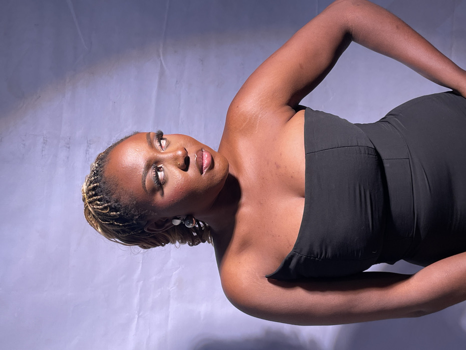 A medium-shot portrait features a single individual, a woman with dark skin, positioned slightly off-center to the left against a light lavender or pale gray backdrop. Her hair is styled in thin, light brown or blonde box braids, pulled up into a high ponytail, with some longer, darker strands and loose curls visible around her right ear. She wears a strapless, form-fitting black garment, likely a dress, with a visible seam running vertically down the bodice.
The woman's head is turned to her right, and her gaze is directed upwards and to the right, beyond the frame. Her lips are slightly parted. She has prominent eyelashes, possibly false, and visible eye makeup. A subtle shine is apparent on her nose and upper lip. On her visible right ear, she wears an earring composed of two spherical elements, one larger and darker beneath a smaller, lighter one. Her right arm is bent at the elbow, with her hand resting on her hip, though the hand itself is not fully visible within the frame. The left shoulder and upper arm are visible.
The backdrop appears to be a fabric sheet, exhibiting visible creases and wrinkles. Lighting is focused on the subject, creating a subtle shadow on the right side of her face and body, as well as on the backdrop directly behind her right side. The ambient lighting casts a cool, slightly blue-purple hue on the backdrop. There are no discernible elements in the image to indicate a specific geographic location within the city of Jos, Nigeria, or any city.