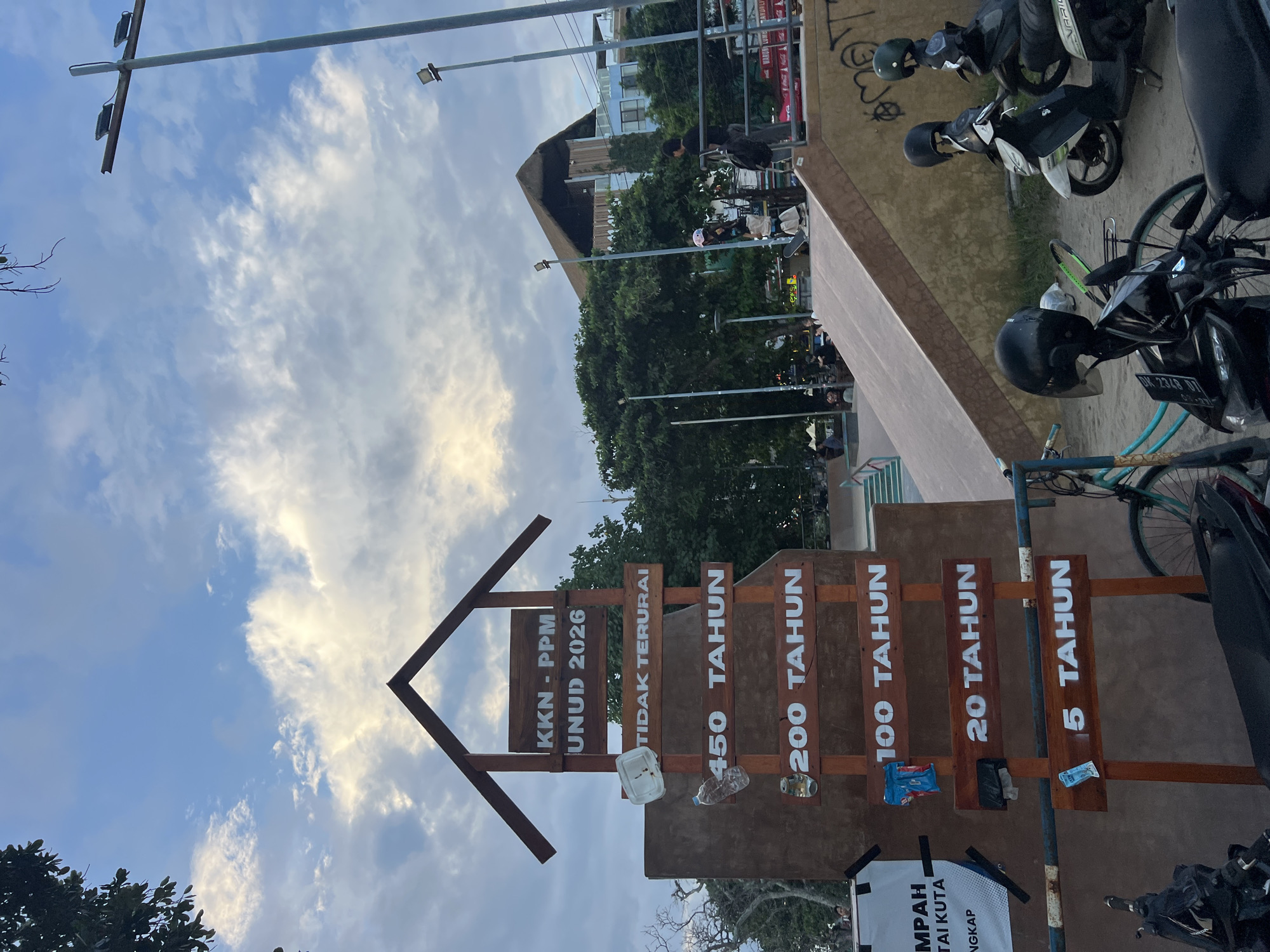 The image, taken in the city of Kuta, Indonesia, presents a sign with an arrow pointing upwards, labeled "KKN-PPM UNUD 2026". Below the arrow, the sign features a series of wooden planks displaying time periods: "TIDAK TERURAI", "450 TAHUN", "200 TAHUN", "100 TAHUN", "20 TAHUN", and "5 TAHUN". Litter, including plastic containers and a water bottle, is attached to the sign. A partial view of a sign with the word "MPAH" and the words "TAI KUTA" is visible at the bottom. The background includes trees, a thatched-roof building, and streetlights against a cloudy sky. Motorbikes are parked on the right side of the frame.