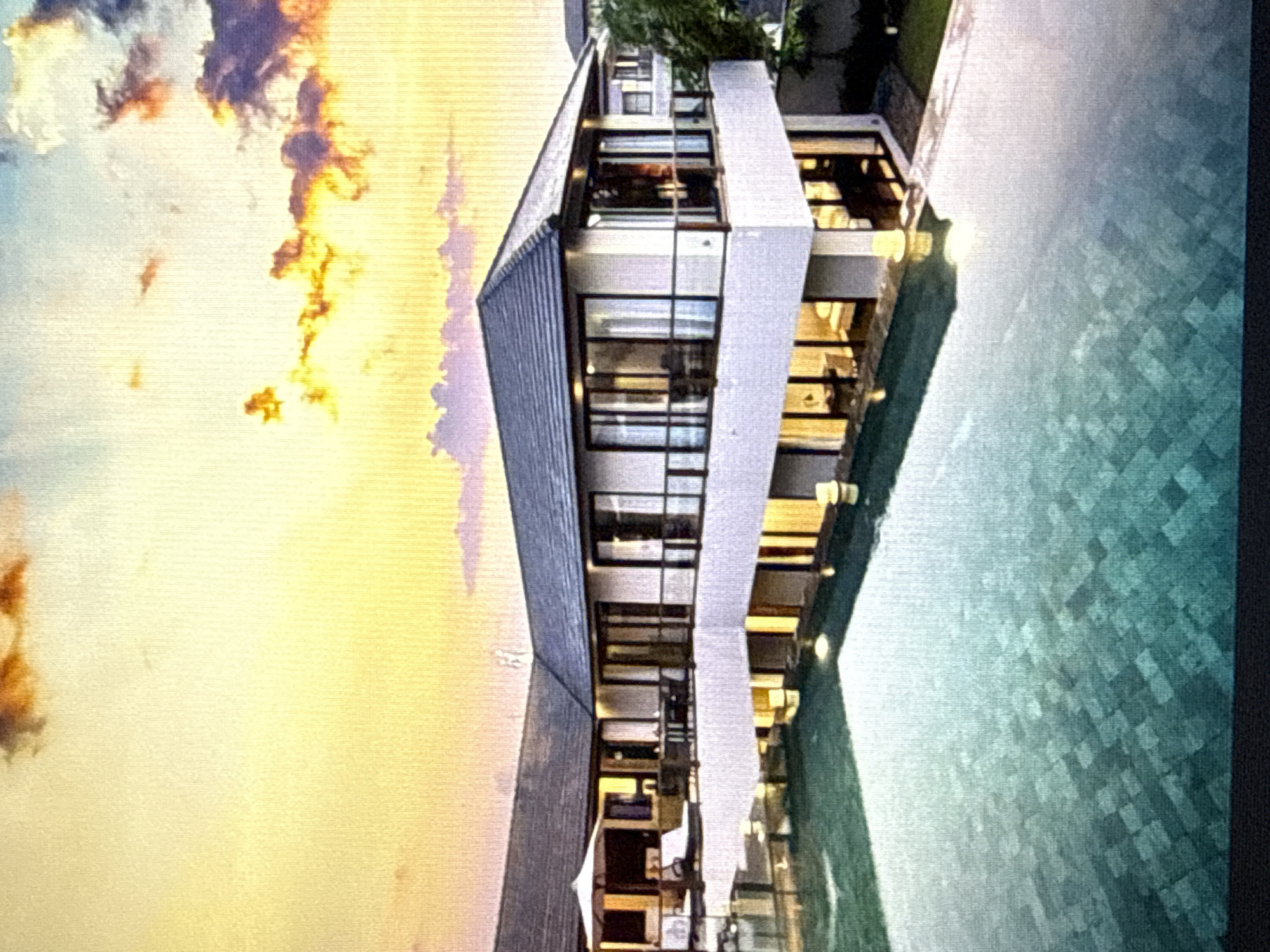 An exterior, eye-level, medium shot captures a luxurious modern villa with an infinity pool at sunset. The villa is multi-story with white walls, dark trim, and a dark, sloped roof. Large glass windows and doors dominate the facade, offering views into brightly lit interior spaces. A balcony or terrace wraps around the upper level, furnished with outdoor seating and a large white umbrella. The infinity pool, tiled in shades of teal and dark green, reflects the warm hues of the sunset and the lights of the villa. The sky is ablaze with orange, yellow, and purple clouds, casting a dramatic glow over the scene. The overall impression is one of serene opulence and tranquil relaxation.