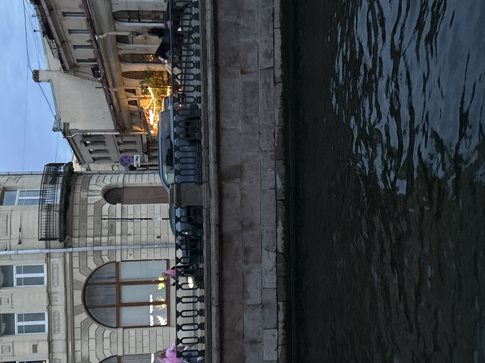 The image shows a view of a canal in an urban setting. On the far bank of the canal, there is a stone wall and a black metal railing. Behind the railing, there are buildings with architectural details. On the left, a building has a large arched window and multiple smaller windows. Two individuals wearing pink clothing are visible on the sidewalk. To the right, other buildings line the street, with some displaying illuminated decorations, possibly for a holiday. A dark-colored car is parked or driving along the street. Further down the street, a horse-drawn carriage is visible. The water in the canal is dark and choppy, reflecting the dim light. The sky above is a dusky blue, suggesting it is either dawn or dusk.