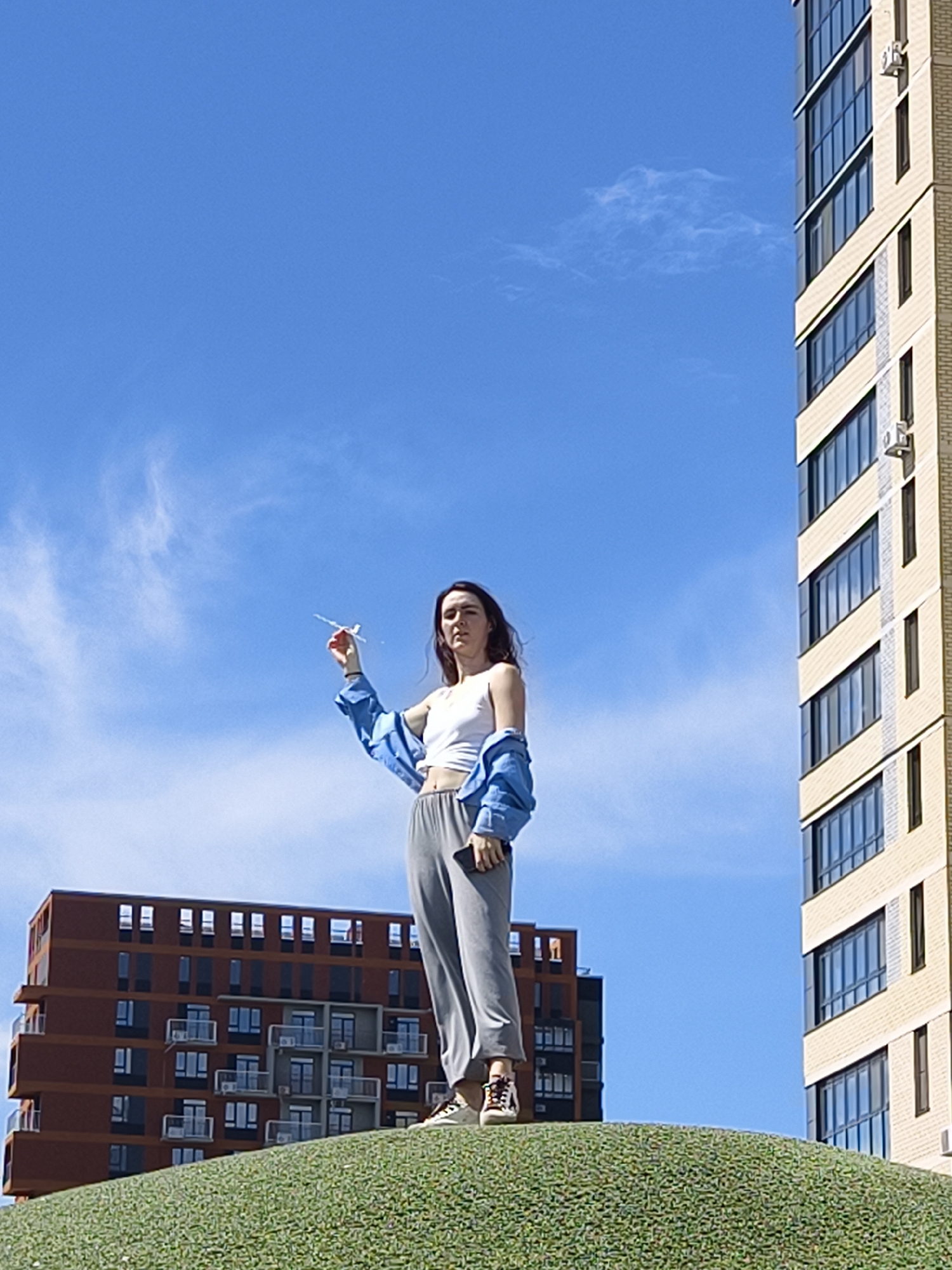 An outdoor, eye-level shot on a bright, sunny day in Tyumen, Russia, captures a young woman standing on a verdant, low-lying mound. She faces slightly towards the left, her head tilted slightly. She holds a small, white model airplane out with her right hand. The woman has long dark hair and is dressed in a white crop top, loose grey trousers, and a light blue unbuttoned shirt or jacket worn over her shoulders. She wears light-colored sneakers. The sky above is a clear, vibrant blue with minimal wispy clouds. In the background, two modern high-rise buildings frame the scene. On the left, a multi-story building features reddish-brown facades with numerous windows and balconies. To the right, a taller, lighter-colored (possibly beige or light yellow) building dominates the right edge of the frame, showcasing many reflective windows and external air conditioning units. No discernible text is visible on any objects or surfaces.