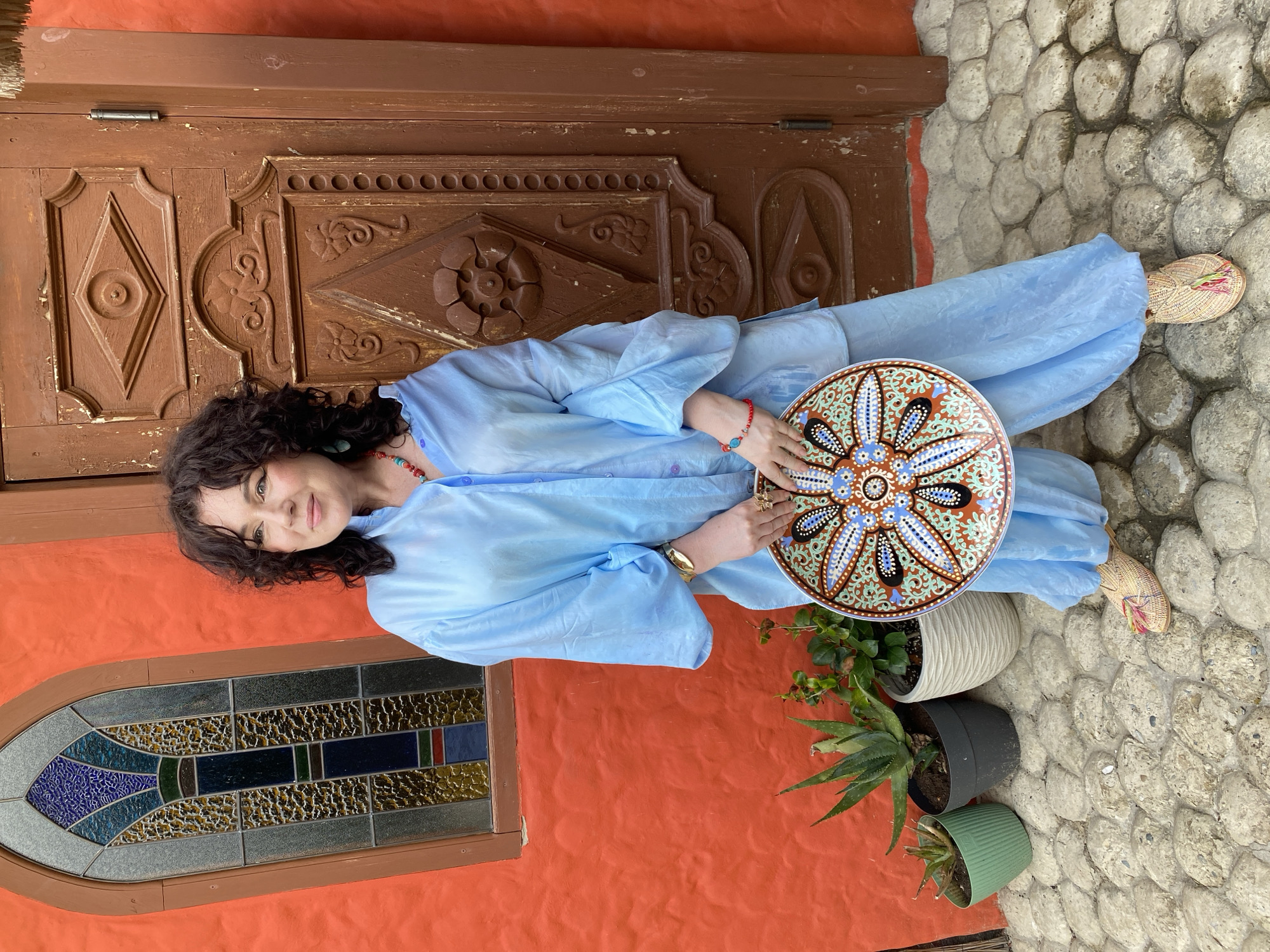A woman with curly brown hair and a friendly expression is holding a decorative plate with intricate patterns. She is wearing a flowing, light blue garment. The background features a rustic wooden door with carved details, an orange textured wall, and a stained-glass window. Several potted plants, including an aloe vera plant, are visible. The ground is covered in large, irregularly shaped stones. To the right, a small woven fan is placed on the stone ground. The scene appears to be outdoors, possibly in a courtyard or near a building entrance. The lighting suggests it could be daytime. The overall impression is one of relaxed charm.