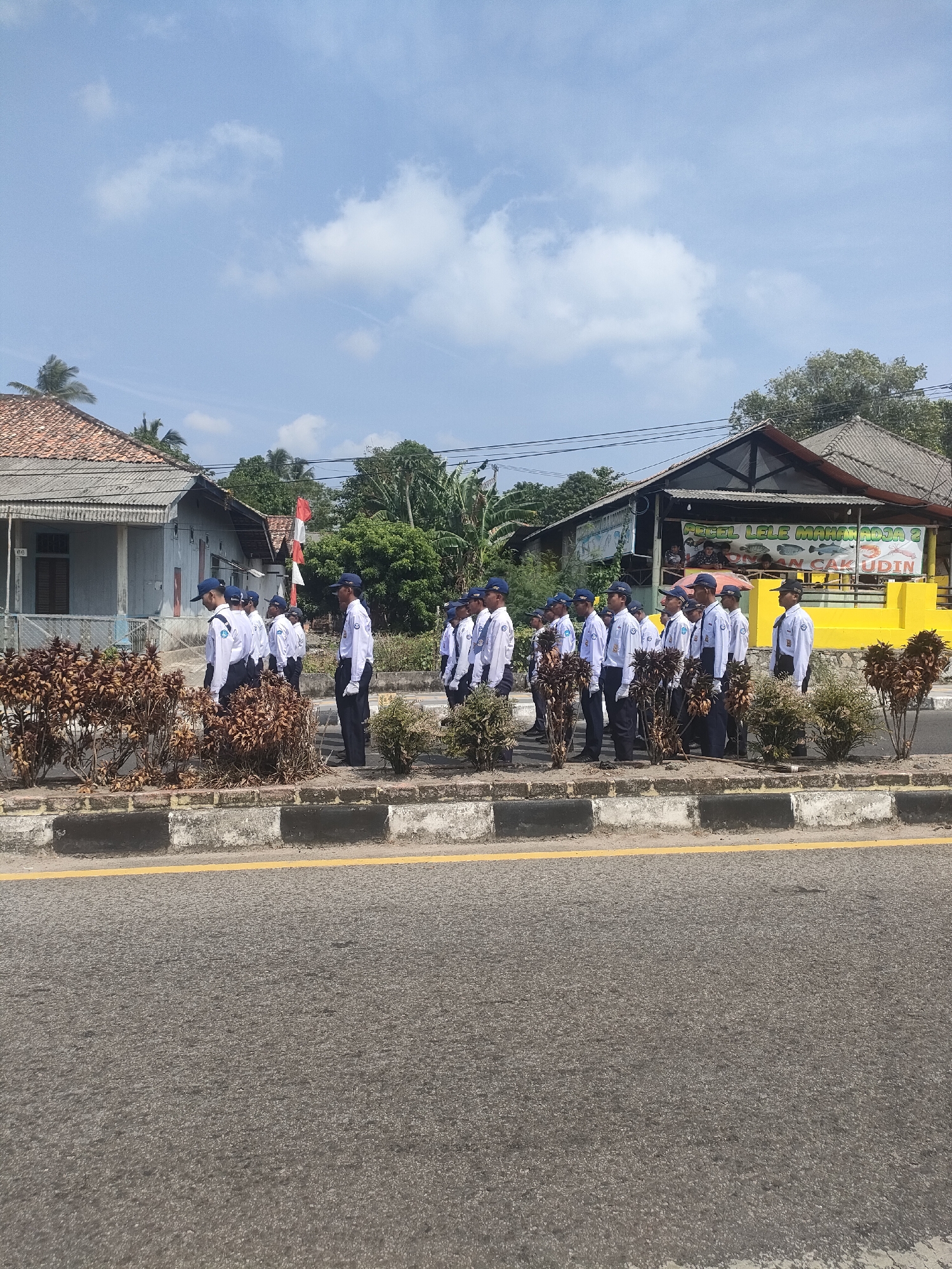 In Sungailiat, Indonesia, a group of young men dressed in white shirts, dark pants, and blue caps stand in formation along a sidewalk. They appear to be participating in an outdoor event or ceremony, as evidenced by their neat attire and organized arrangement. Behind them, a red and white Indonesian flag is partially visible, fluttering in the breeze. The background features a street with a yellow line marking, bordered by a concrete curb. Residential buildings with tiled roofs and a sign that reads "PECEL LELE MAHRUS ROJO 2" are also present. The sky above is a clear blue with scattered white clouds, suggesting a sunny day. The vegetation along the sidewalk is dry and brown, contrasting with the lush green trees further in the background.