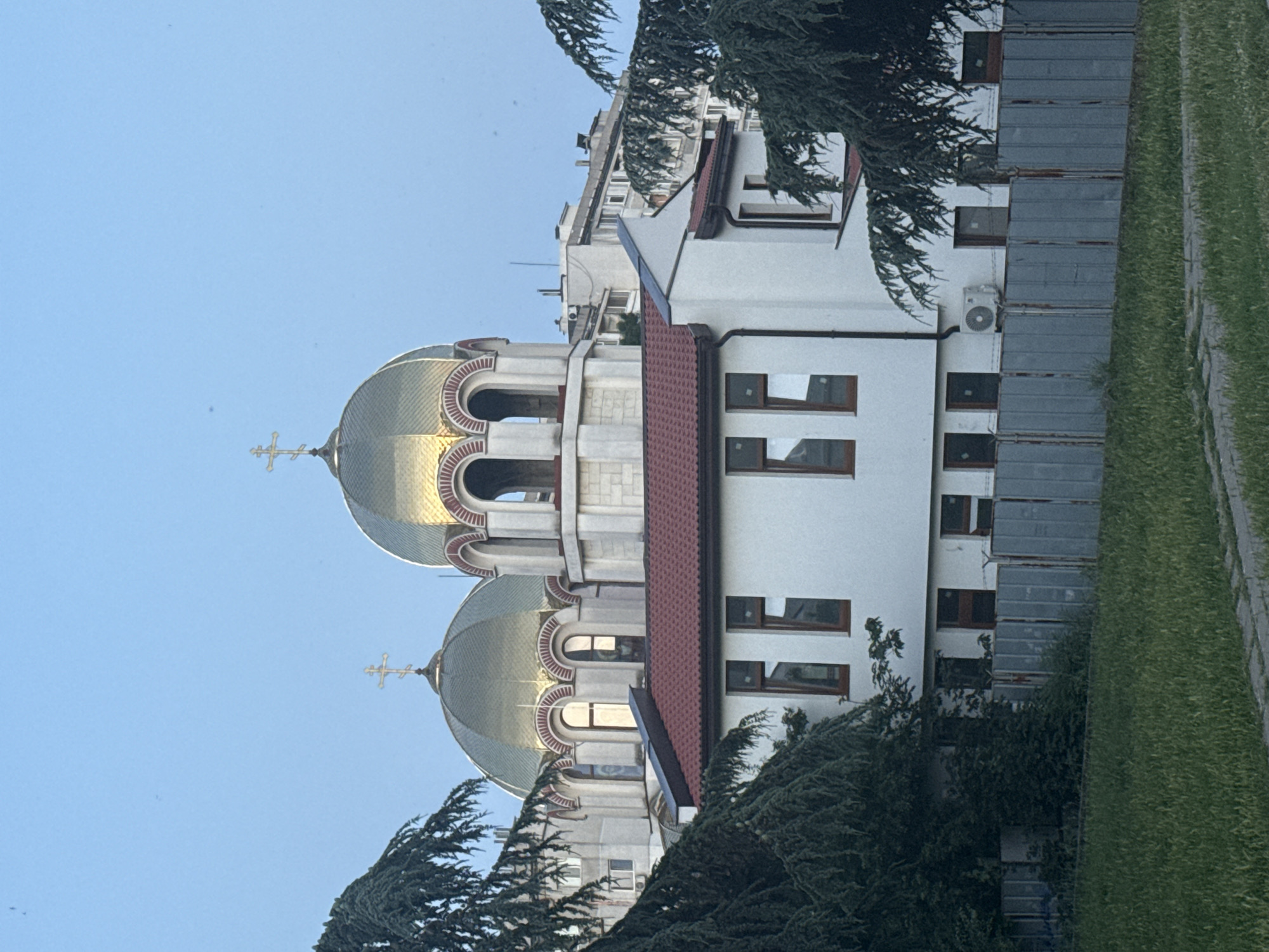 The image shows the exterior of the St. Nicholas the Wonderworker Cathedral in Burgas, Bulgaria, on a clear day with a light blue sky. The cathedral features prominent golden domes topped with crosses. In the foreground, a white building with a red tiled roof is visible, with several windows. Trees with dark green foliage partially obscure the view of the cathedral and the white building. A grey metal fence runs horizontally across the bottom of the image, separating the buildings from a grassy area in front of it. Behind the cathedral, other buildings of varying heights and architectural styles can be seen. There are no people or animals visible in the image. The scene appears to be a quiet, daytime view of the religious landmark.