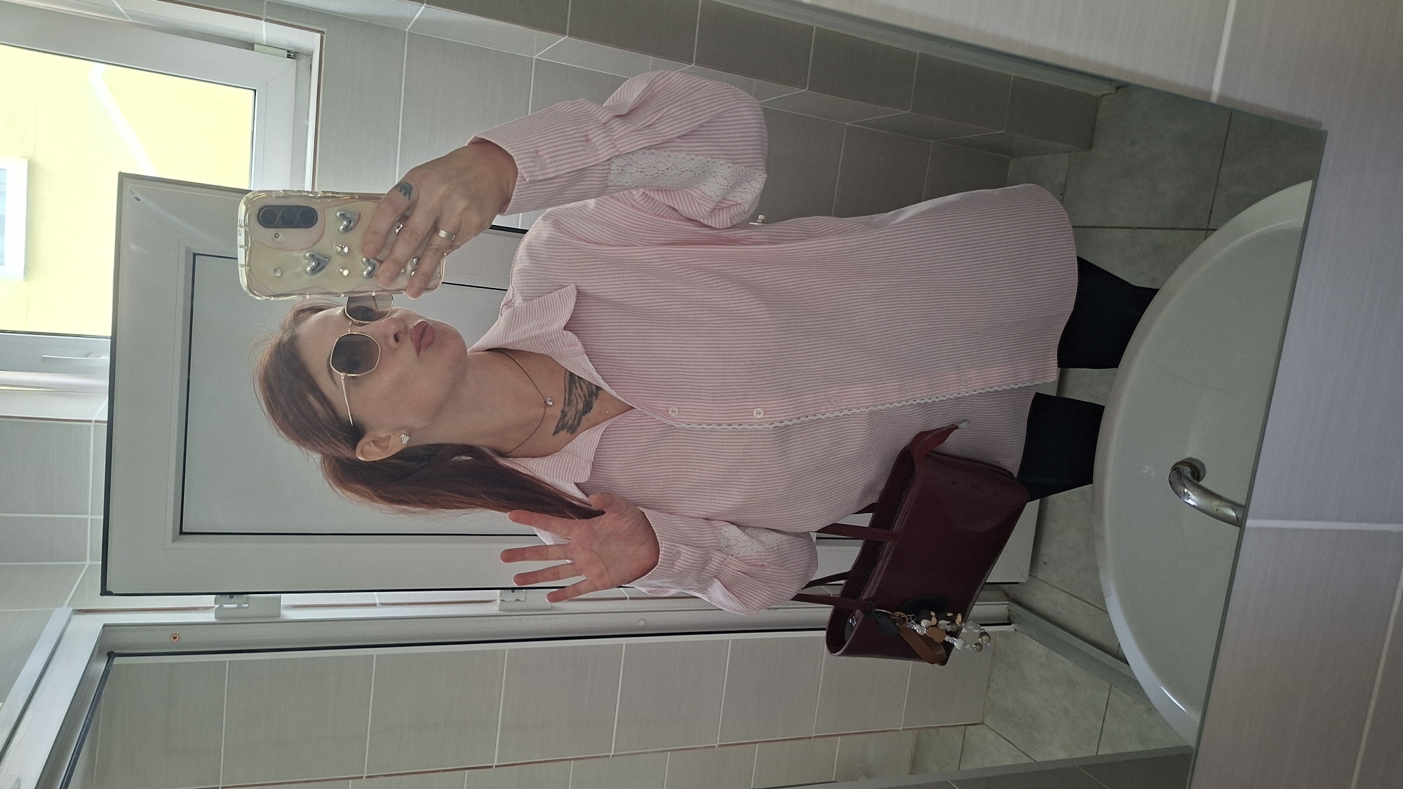A woman with auburn hair in a ponytail is reflected in a bathroom mirror, taking a selfie. She is wearing a pink and white striped button-down shirt, black leggings, and sunglasses. She has a burgundy handbag with charms hanging from it. Her left hand is raised in a waving gesture. The bathroom features tiled walls and a sink in the foreground. The reflection of a yellow window is visible in the background. The image appears to be taken in a residential bathroom. The exact location within Myadzyel, Belarus cannot be determined from the visual information provided.