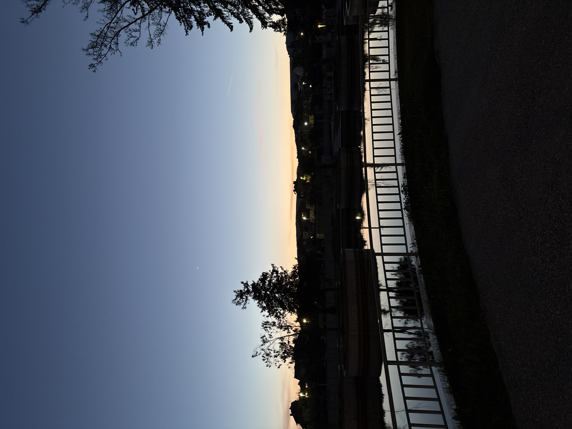 The image depicts a twilight scene in Panagyurishte, Bulgaria. The sky transitions from deep blue to orange near the horizon. A dark silhouette of trees is visible against the sky, along with faint city lights. A metal railing runs along the right side of a body of water, reflecting the sky and lights. To the right of the railing is a dark path or road. The image is taken at an angle, with the horizon appearing vertical.