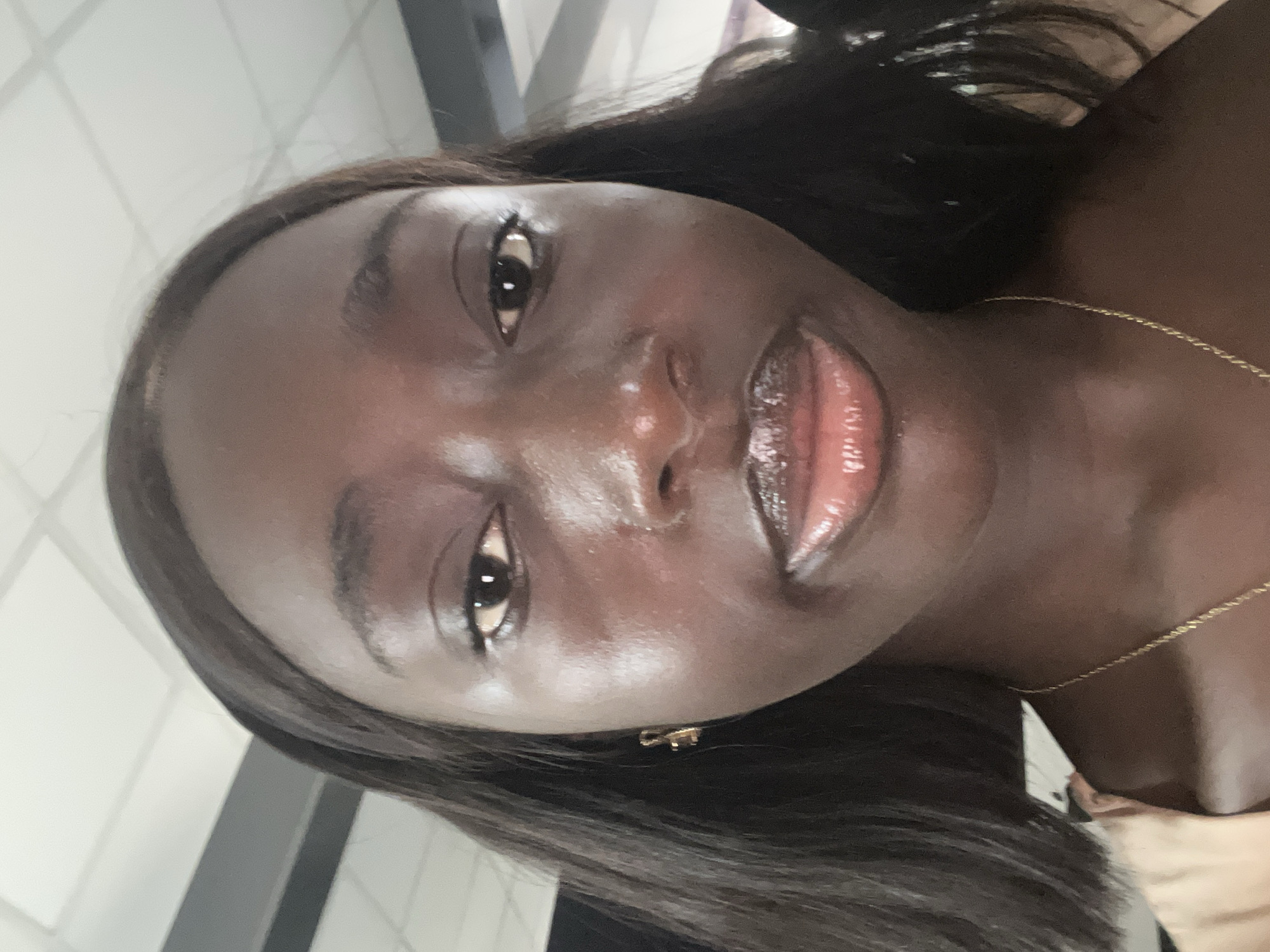 A close-up selfie of a dark-skinned woman with long, dark hair. She is wearing a gold necklace and small gold earrings. Her skin appears glossy, and her lips are shiny with gloss. No context to indicate the location.