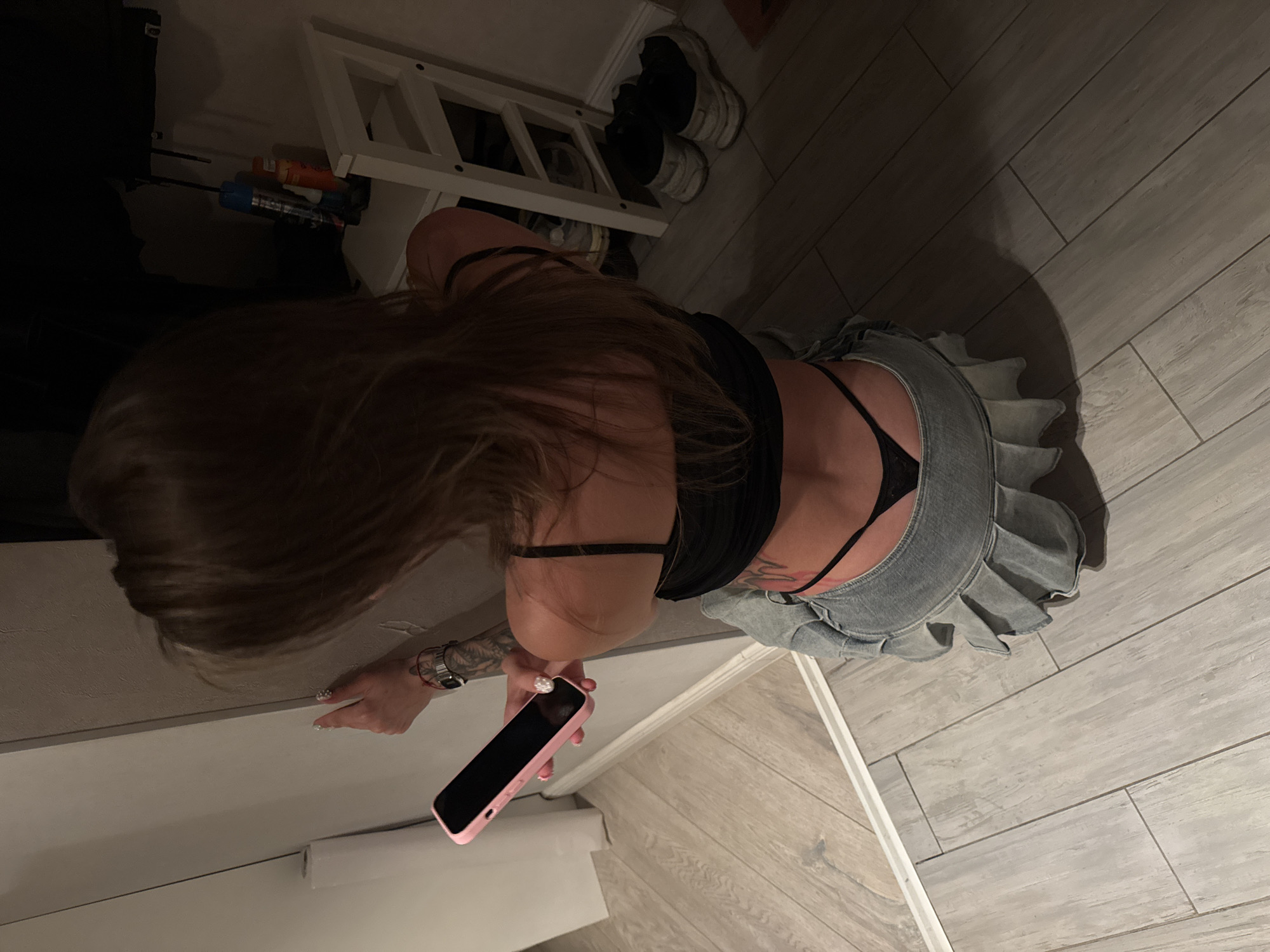 The image shows a woman from the back, kneeling on a wooden floor. She has long, dark brown hair and is wearing a black top and a denim skirt with ruffles. A black thong is visible. She is holding a pink smartphone in her right hand.
To her left, there is a shelf unit with various items, including what appear to be spray cans, one blue and one orange. Behind the shelf unit, a white ladder-like structure is visible, with a pair of white sneakers placed on one of its shelves. The overall lighting suggests it is nighttime or indoors with dim lighting.
The setting appears to be a room in a residential building, possibly a hallway or a room with utilitarian storage. The floor is tiled with a wood-grain pattern. There are no visible emotional cues from the subject due to her pose. No text is visible in the image. The location context provided is Moscow, Russia.