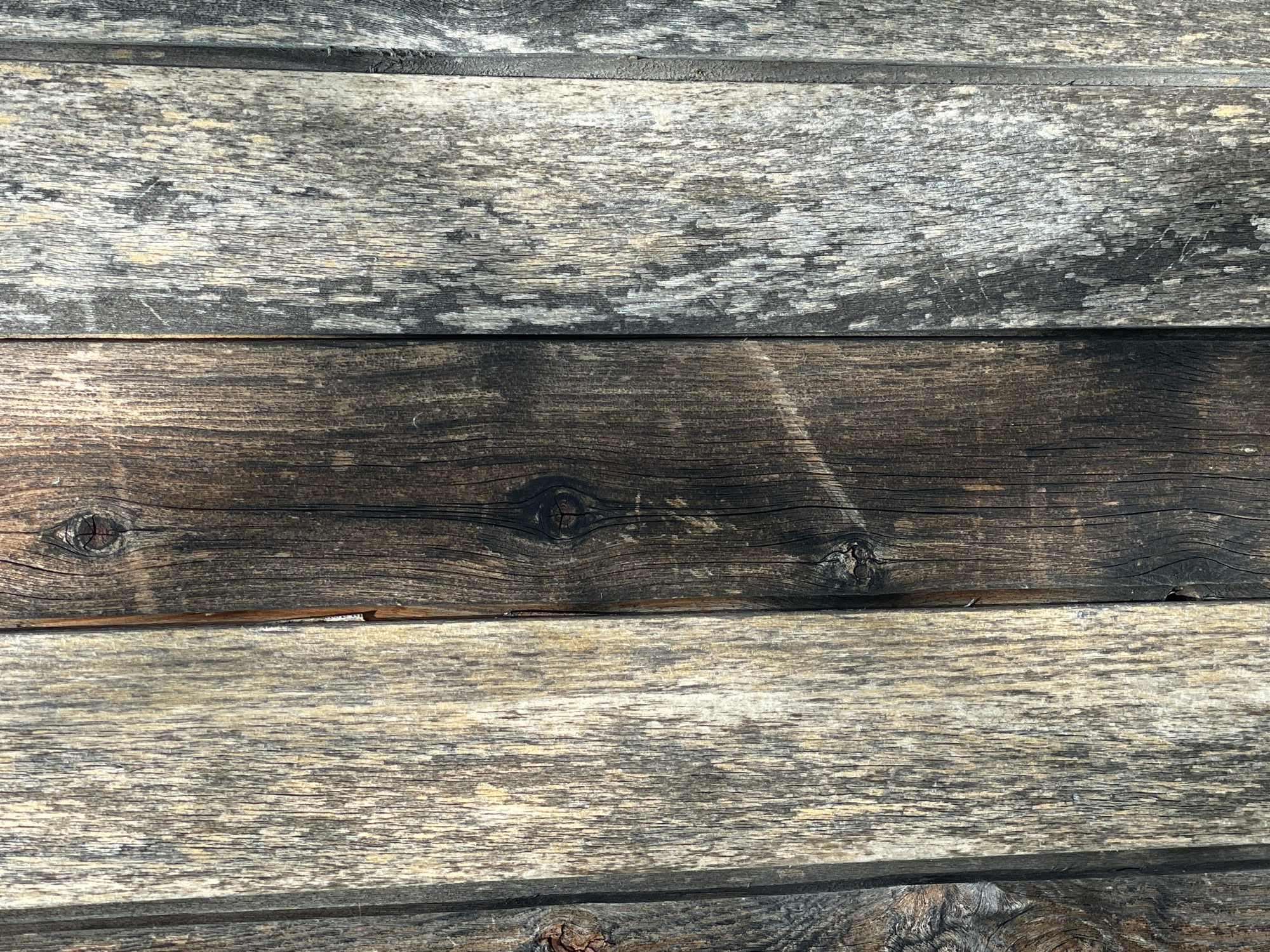 The image is a close-up, vertical, full-frame shot of a weathered wooden wall. The wall is composed of three vertical planks. The leftmost plank is a light, weathered gray with visible grain and some darker vertical streaks. The middle plank is significantly darker, a rich brown with black charred-looking areas, particularly around a prominent knot near the center. The rightmost plank is also weathered gray, similar to the leftmost plank, but appears to have more patches of lighter color and a rougher texture. The planks are joined vertically, with slight gaps and overlaps creating subtle shadows. The lighting suggests a natural light source, possibly sunlight, casting faint shadows and highlighting the textures of the wood. There are no people, animals, or other objects visible in the image. The overall impression is one of age and outdoor exposure.