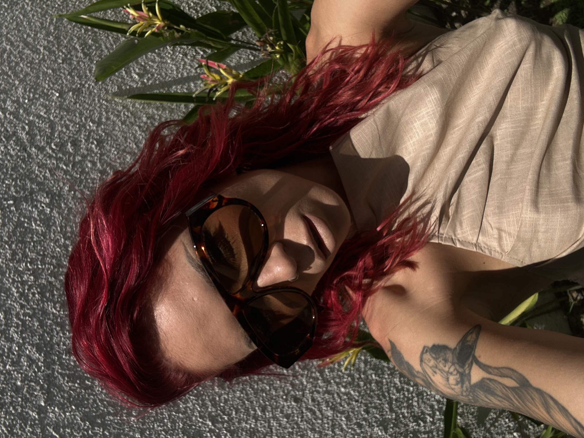 The image shows a person lying on a textured gray surface outdoors. The person has red hair, is wearing sunglasses, and a light brown linen shirt. There is a floral tattoo on their left arm. Green leafy plants with yellow and pink flowers are scattered around the person's head and body. The location is possibly North Kuta, Indonesia.