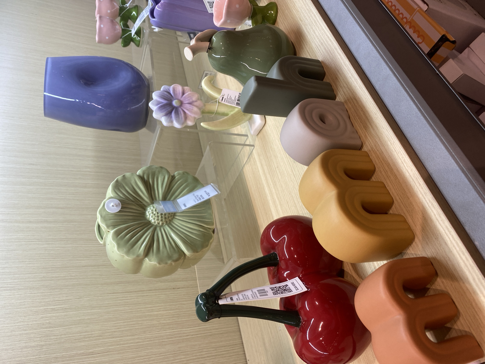 The image shows a retail display shelf in Denpasar, Indonesia, featuring various decorative ceramic and glass items. On the top shelf, there is a light green ceramic object shaped like a flower, a transparent purple vase with a hole in the center, and two pink ceramic flower decorations. Below, on a lower shelf, are a pair of glossy red ceramic cherries, a pastel purple ceramic flower on a stem, a green ceramic pear, and a pink ceramic cup with a stem. In the foreground, on a wooden shelf, are several matte ceramic objects in abstract shapes and colors: two orange arches, a beige rolled shape, and a dark green chair-like object. The background is a light wood-paneled wall. Tags are visible on several of the items, including QR codes.