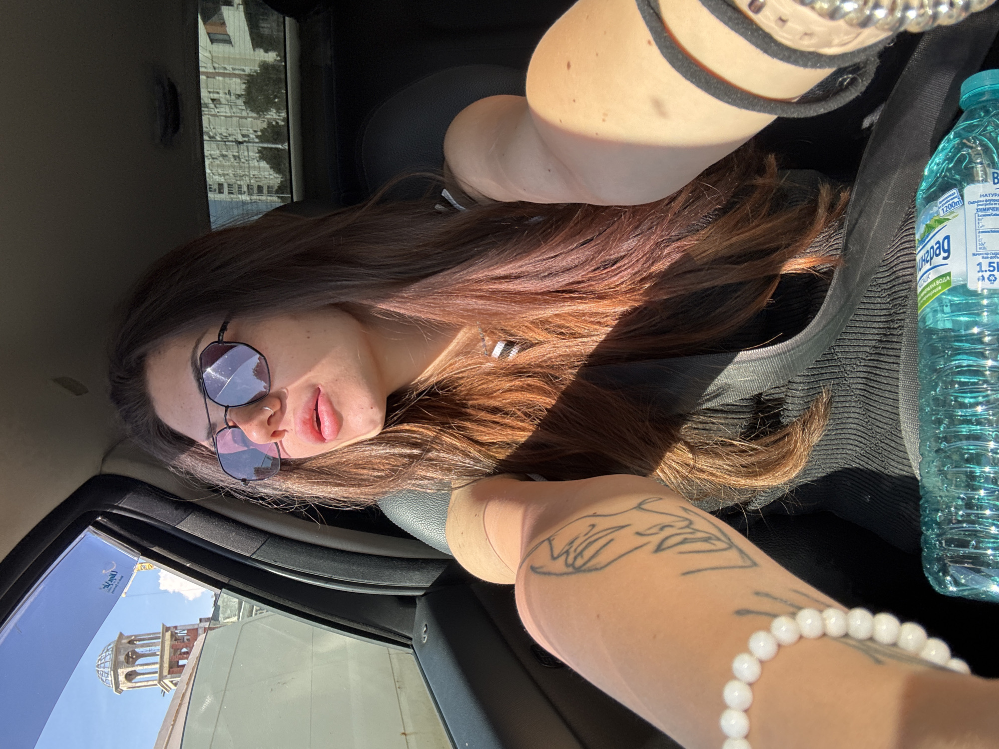 In Sofia, Bulgaria, a young woman with long, reddish-brown hair is seen from the chest up, sitting in the driver's seat of a car. She is wearing sunglasses and looking to her left, out the window. Her left arm is extended, holding the phone, and her right arm is resting on the steering wheel. A tattoo is visible on her left forearm. A white beaded bracelet adorns her left wrist. In the background, through the car windows, a domed building and residential buildings are visible, suggesting an urban setting. A bottle of water is placed on the console in front of her.