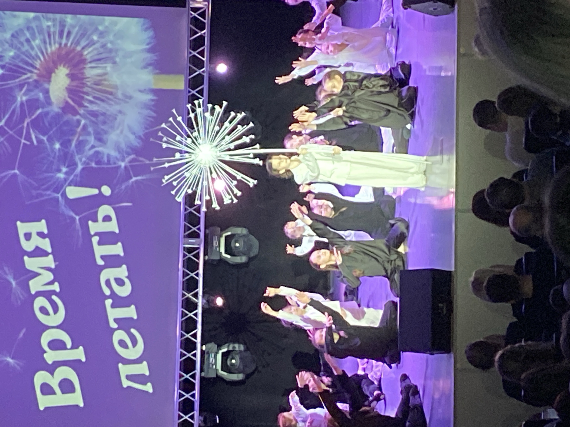 Here is a description of the image:
The image shows a stage performance likely in Chaikovsky, Russia, based on context. A screen in the background displays a dandelion image and the text "Время летать!" (Time to fly!). A decorative dandelion sculpture stands on the stage. Several performers are on stage; some are dressed in white robes while others are in black robes. They appear to be engaged in a dance or theatrical performance. A partial view of the audience is visible on the right. Stage lighting fixtures are also visible.