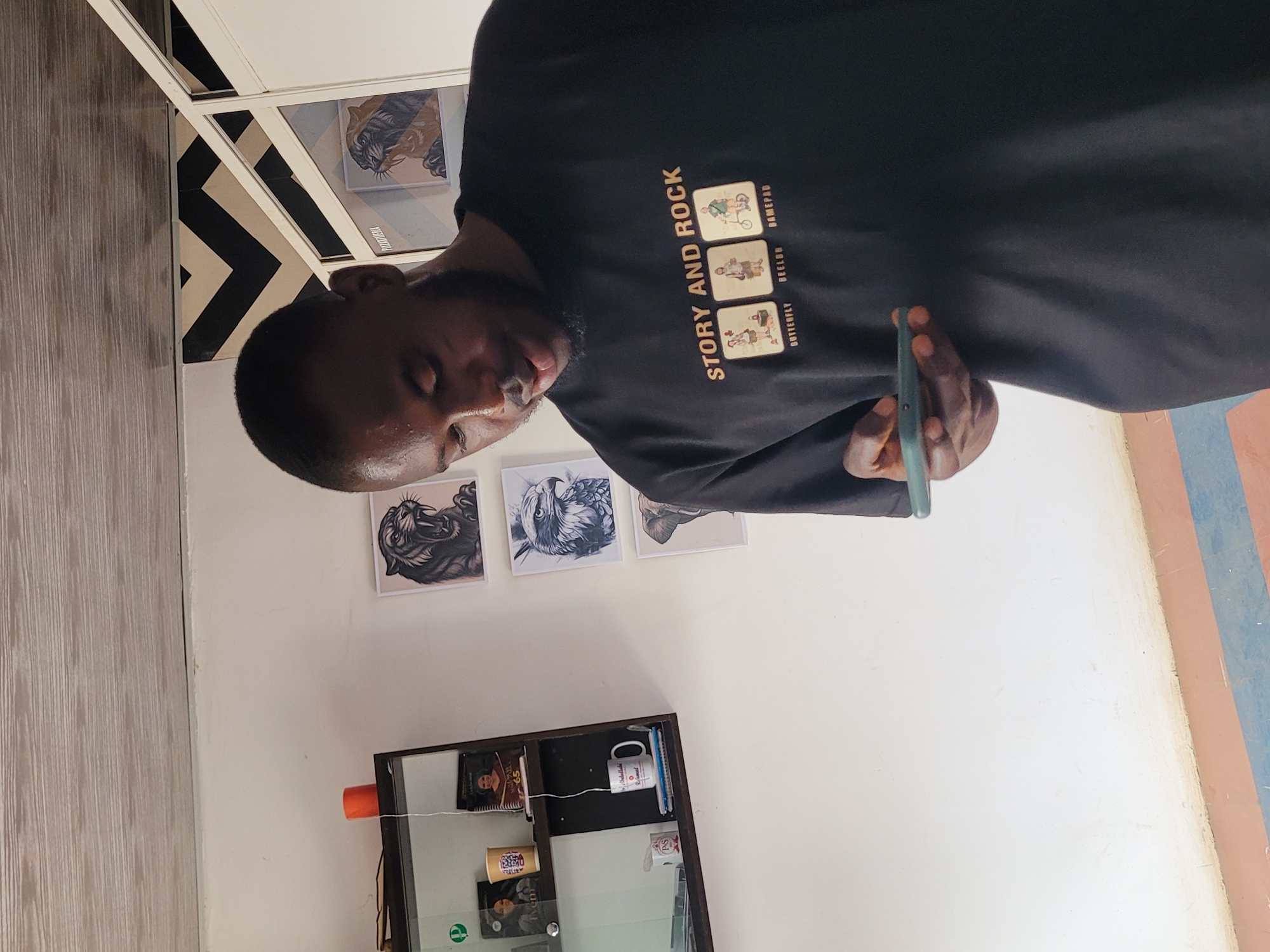A man is holding a mobile phone in an indoor setting, likely in Jos, Nigeria. He is wearing a black t-shirt with the words "STORY AND ROCK" printed on it. On the wall behind him are several framed images depicting animals, possibly tigers and owls. There is a glass-fronted cabinet with various items inside, including books and a mug. The floor has a light-colored, wood-like finish, and there is a geometric design on a wall or structure near the top left corner. The wall near the floor has vertical red and blue stripes.