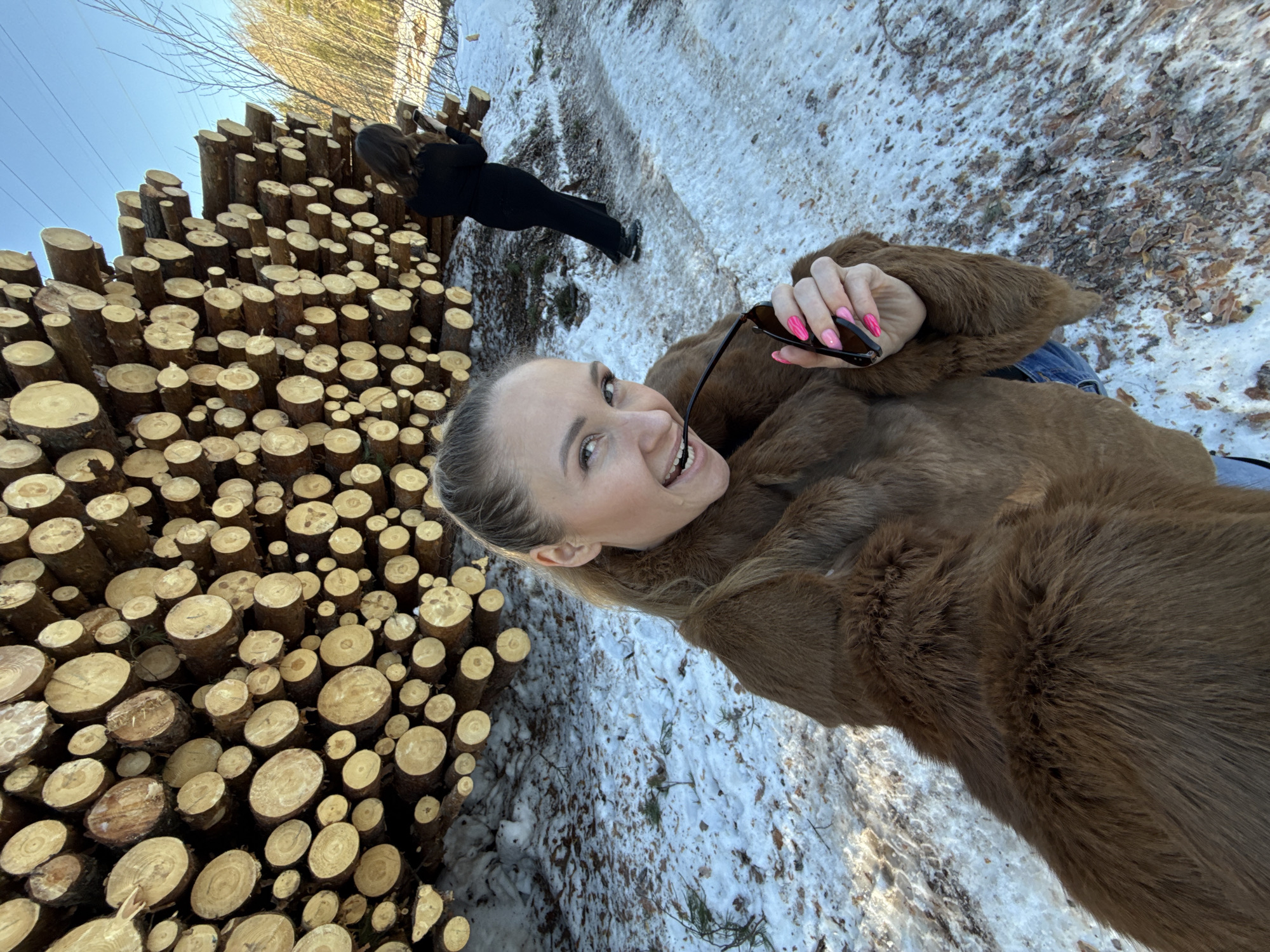 The scene depicts two women outdoors in a snowy, forested area in Noviy, Russia. In the foreground, a blonde woman with hair pulled back, wearing a brown faux fur coat and blue jeans, is smiling broadly at the camera. She holds dark sunglasses in her right hand, with one arm of the glasses resting in her mouth. Her fingernails are painted bright pink. Behind her, a massive stack of cut logs forms a prominent feature across the midground, with various diameters of light-colored wood cross-sections visible. Further back and to the right, a second woman with dark hair, dressed in a long-sleeved black top and black pants, stands on a partially snow-covered path, facing away from the camera towards the stacked logs and a sparse forest with bare trees. The ground throughout the scene shows patches of snow, ice, and brown leaf litter. A clear blue sky with faint power lines is visible in the upper right portion of the background.