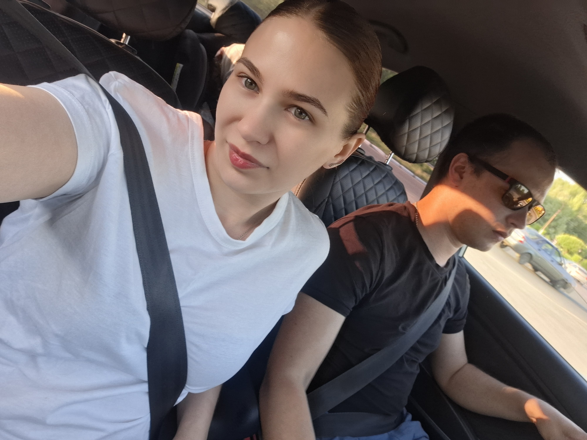 A woman and a man are sitting inside a car wearing seatbelts. The woman has fair skin, her hair is pulled back, and she is wearing a white t-shirt. The man is wearing sunglasses and a black t-shirt. Outside the car, there is a parking lot with cars and trees. The location is Chaikovsky, Russia.