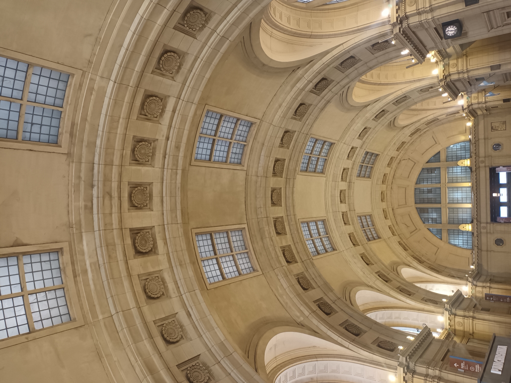 The image captures the interior architectural detail of Galerías Pacífico, a shopping center located in Buenos Aires, Argentina. The view is primarily focused upwards towards the ornate ceiling, which features a series of concentric arches adorned with decorative medallions and evenly spaced windows. The walls are constructed of pale stone. The angle of the shot creates a sense of depth and perspective, emphasizing the curved shapes. Portions of the building's supporting structures, including arches, pillars, and lights, are visible towards the right side of the frame. There are no people visible.