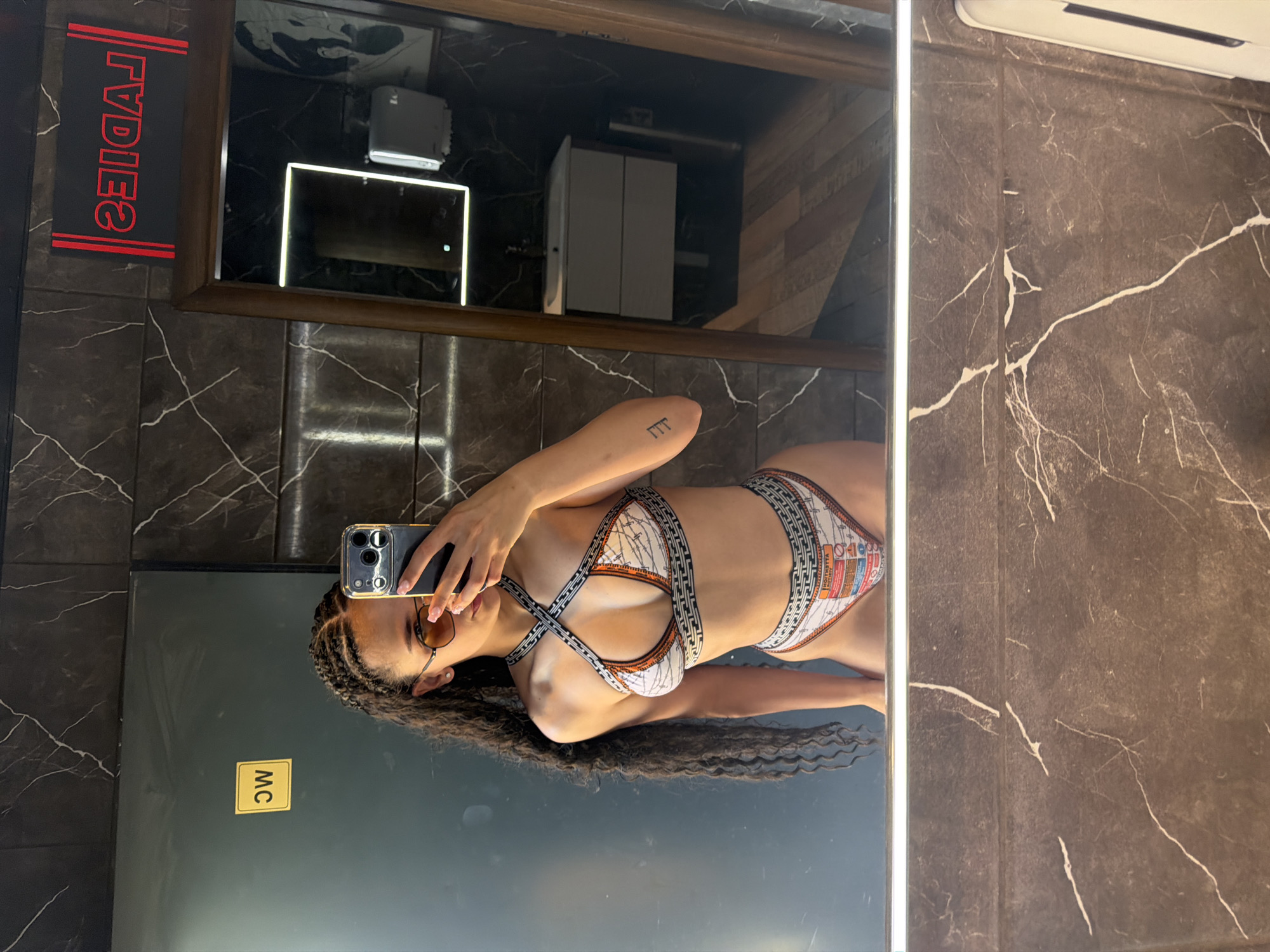 A young woman with braided hair and sunglasses takes a mirror selfie in a restroom. She is wearing a patterned bikini and standing in front of a dark tiled wall and a large mirror. The mirror reflects the woman, a sink and vanity area, and a neon "LADIES" sign. The overall scene appears to be in a modern establishment, potentially a hotel or a public restroom. The city of Nesebar, Bulgaria, is not visible or indicated in this image.