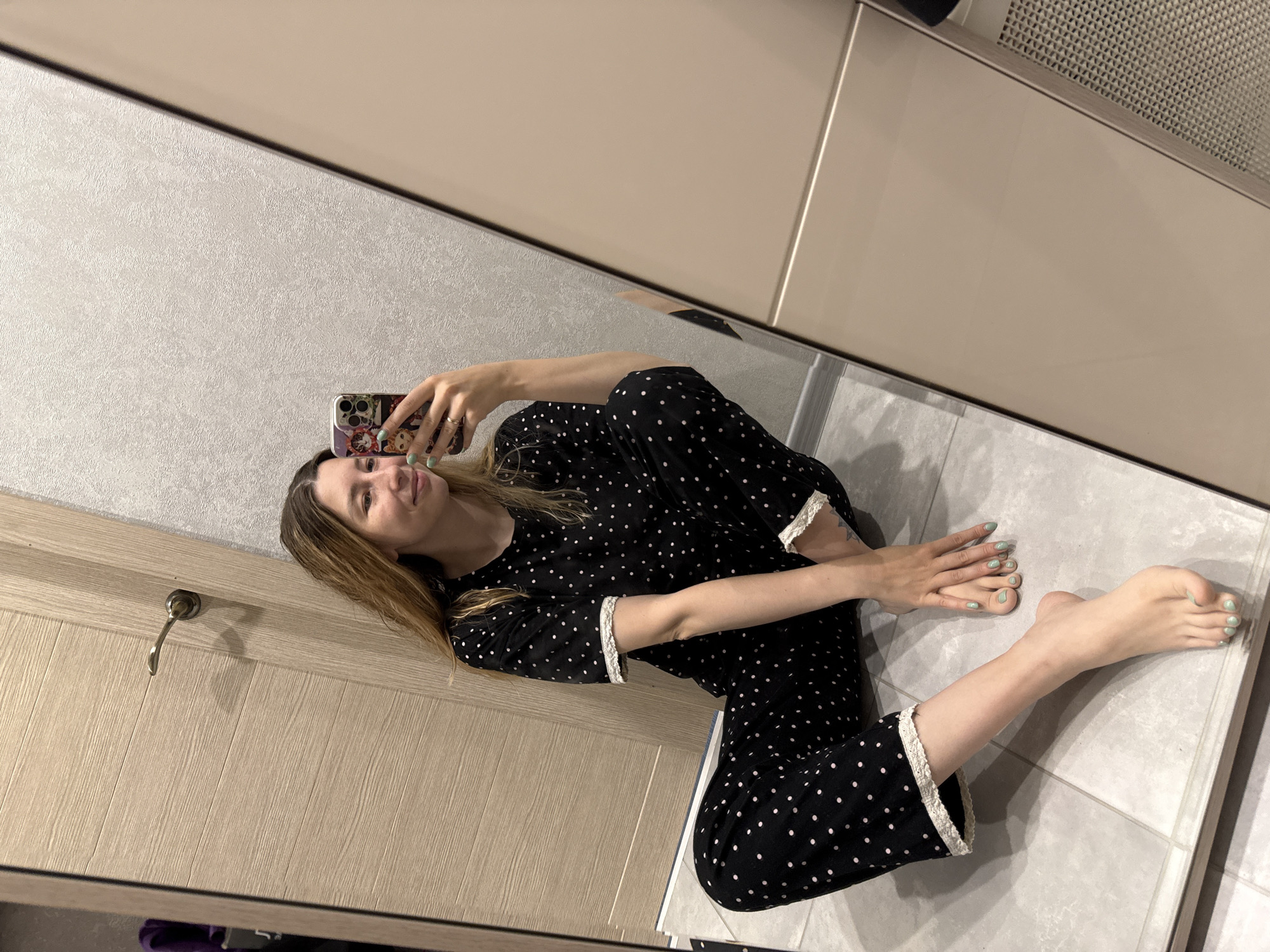 A young woman is taking a selfie in what appears to be a bathroom. She is wearing black pajamas with white polka dots and white trim. She is seated on a gray tiled floor, with her legs outstretched and her feet pointed towards the bottom right of the frame. Her toenails and fingernails are painted a light green color.
The image is a reflection from a large mirror. Behind the mirror, on the left side of the frame, is a light wooden door with a brass handle. Above the mirror is a cream-colored wall or cabinet. The location is likely a bathroom within a residential building, although the city of Moscow, Russia, cannot be confirmed from this image alone.
