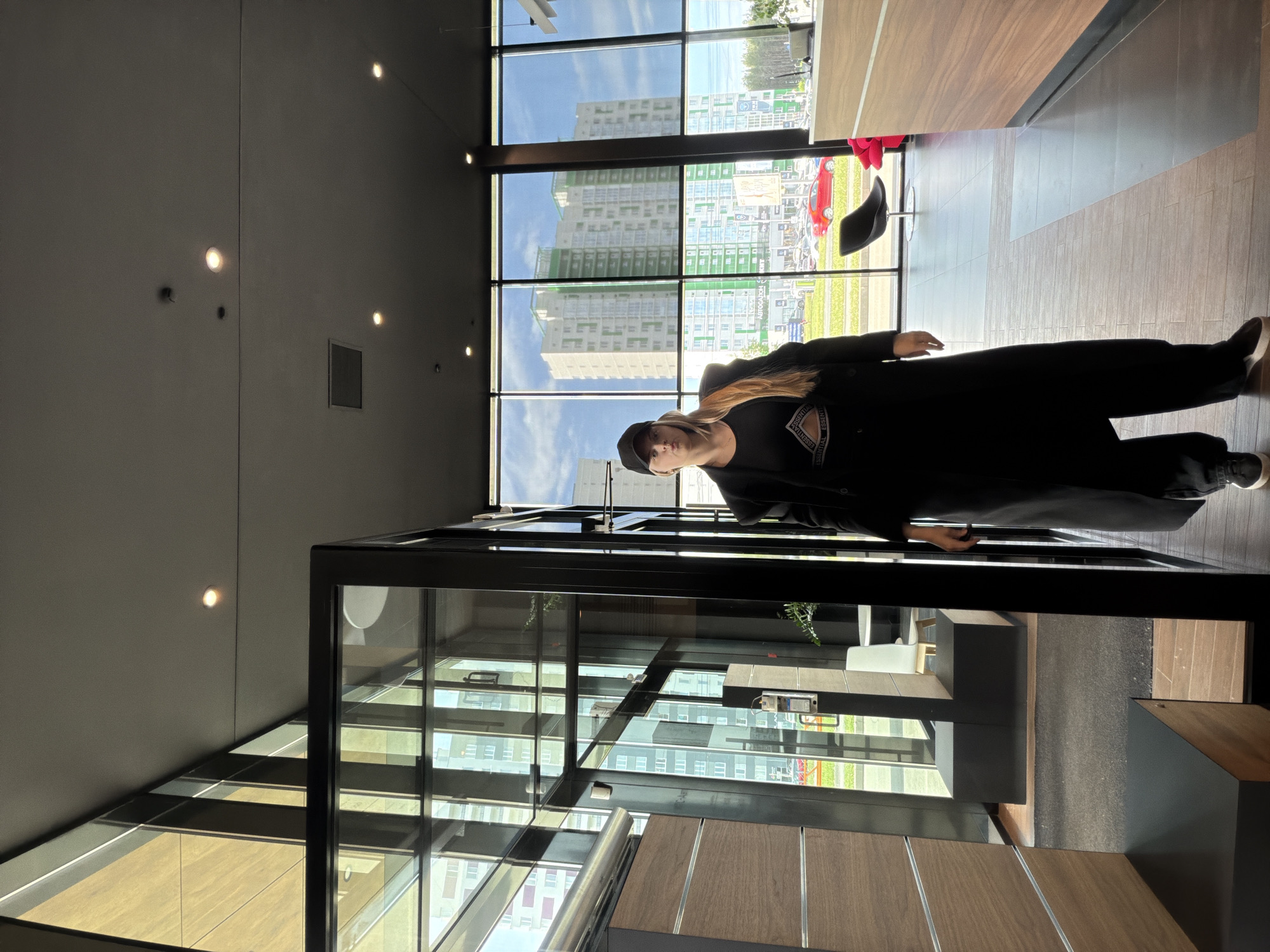 A woman stands in an interior space with a wall of windows displaying a cityscape. The woman is wearing a dark outfit with a baseball cap. She is facing forward. The interior space appears to be a modern lobby or waiting area with wood paneling and glass partitions. Outside the window are multi-story buildings, possibly residential, indicating the location is likely within an urban area. The city is Izhevsk, Russia.