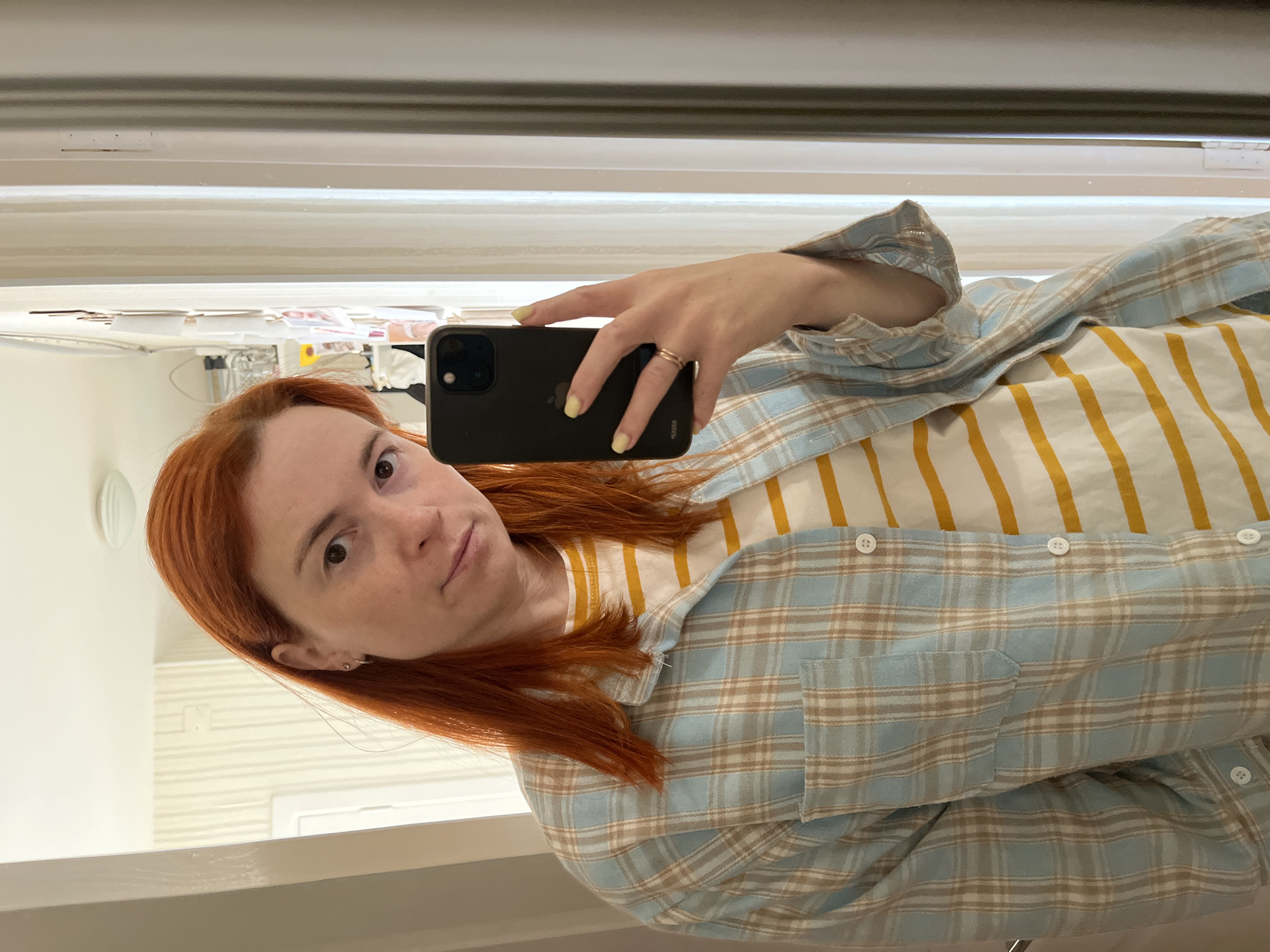 A woman with shoulder-length red hair is taking a mirror selfie in what appears to be a home setting. She is wearing a light blue and tan plaid shirt over a white and yellow striped t-shirt. She has on a ring on her left hand, which is holding a dark-colored smartphone. Her fingernails are painted a light yellow. The background shows a wall with vertical striped wallpaper and a doorway leading to another room. There are some items affixed to the wall in the background, possibly photographs or notes. The lighting suggests it is daytime. The provided context indicates the location is in Chaikovsky, Russia.
