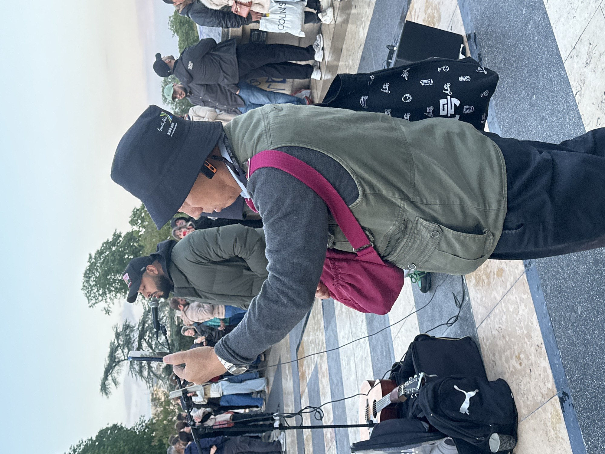 A street musician is positioned in front of a crowd on a patterned stone surface in an outdoor location. The musician is wearing a bucket hat, a gray sweater, and an olive-green vest. A maroon bag hangs over his shoulder. He is holding a smartphone in his hand. A microphone stand and guitar case are at his feet. Another musician, standing in front of a microphone, is visible in the background. Other people are in the crowd, including some holding shopping bags. The sky is overcast. Based on the style of architecture and public space, the location may be within the city of Paris, France.