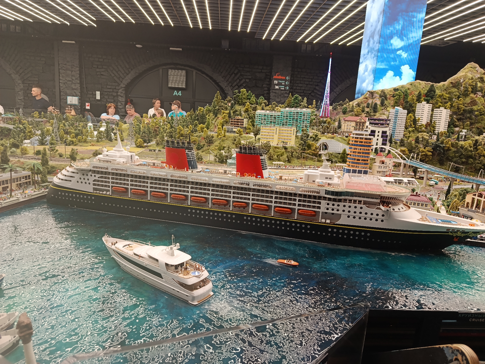 This is a detailed miniature diorama, likely housed within the Grand Maket Rossiya, a prominent model of Russia in Saint Petersburg. The diorama depicts a harbor scene with several vessels. A large cruise ship with a dark hull and yellow accents dominates the scene, featuring rows of lifeboats along its sides and distinctive red funnels with black bands. Moored alongside it is a white luxury yacht with several figures visible on its deck. A small orange dinghy is also present in the water. The water itself is rendered with a textured, shimmering effect to simulate waves. In the background, a detailed miniature city is constructed, complete with buildings, trees, roads, and a distinctive tower with colorful lights. Several people are observing the diorama from a raised walkway in the background, their faces visible as they lean over a railing. The overall environment is dimly lit, with rows of bright lights running across the ceiling above the exhibit.