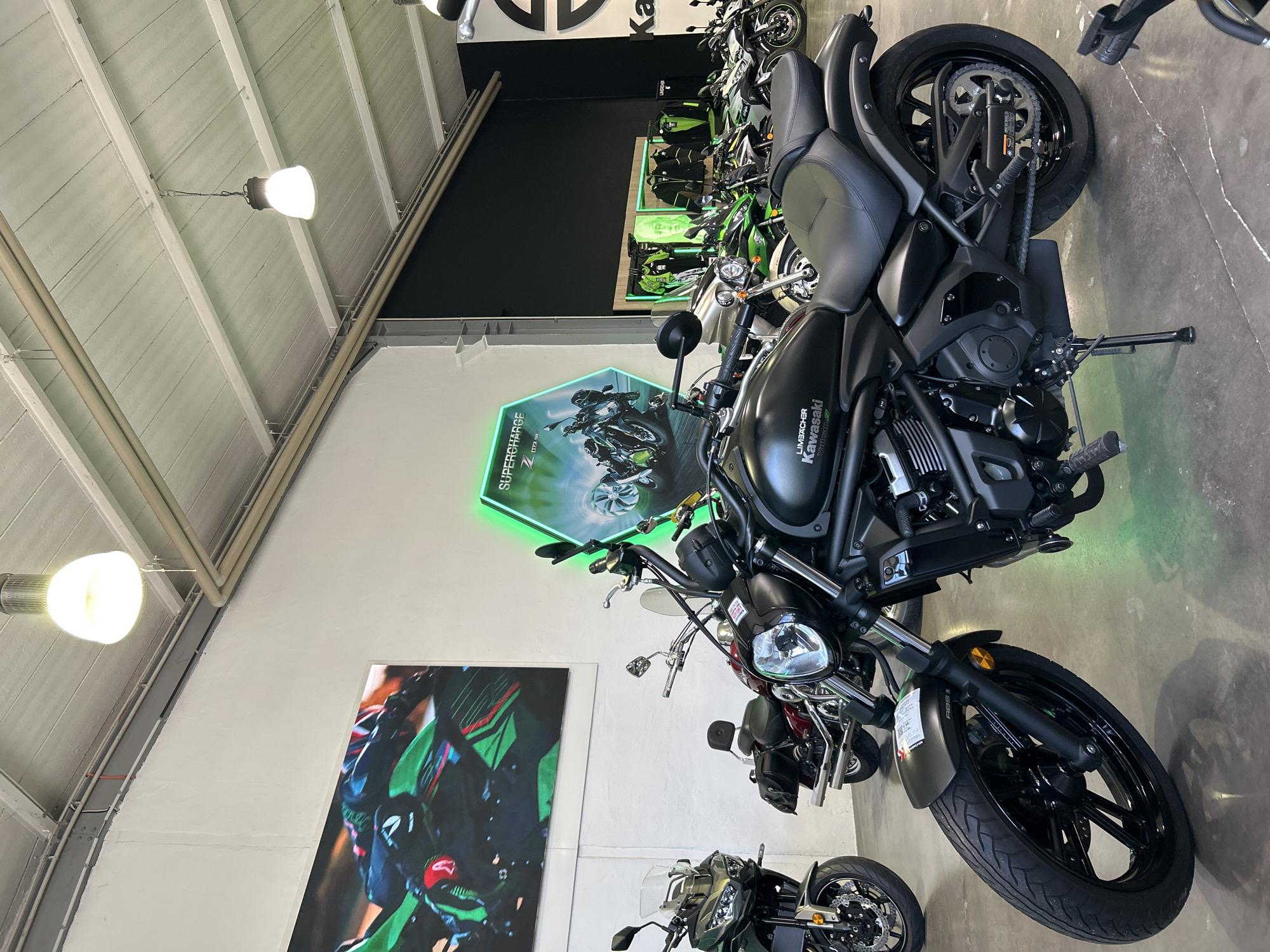 The image depicts the interior of a motorcycle dealership, showcasing various Kawasaki motorcycles. The focus is on a black Kawasaki cruiser-style motorcycle parked in the foreground. Other motorcycles are visible in the background, arranged along the wall. Behind the main motorcycle, there is a hexagonal sign with the word "SUPERCHARGE" and a neon green outline. A large graphic of a Kawasaki motorcycle is on the wall to the left. The ceiling is visible with white beams and several industrial-style light fixtures. The location is in a city of Unknown, Unknown.