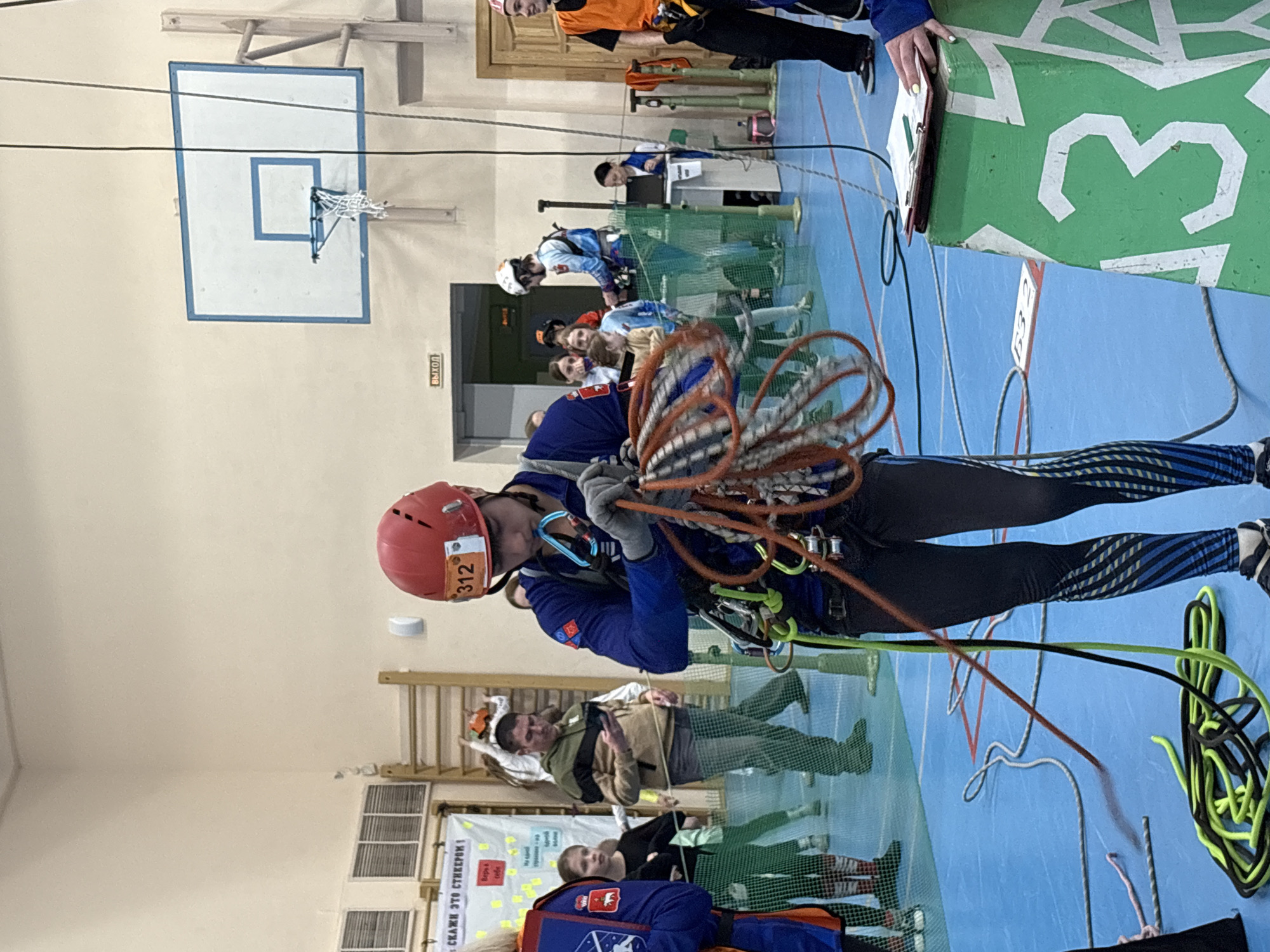 The image shows a man indoors, in what appears to be a gymnasium or sports hall in Berezniki, Russia. He is wearing a red helmet with the number "312" affixed to it, a blue long-sleeved shirt, dark leggings with blue stripes, and climbing gear. He is holding a coiled rope in front of him. In the background, there is a basketball hoop, other people, and various sports-related equipment. The floor is blue, and there is writing in what appears to be Russian on a sign. The number "13" is prominently displayed on a green surface, likely part of the sporting event the image appears to capture.