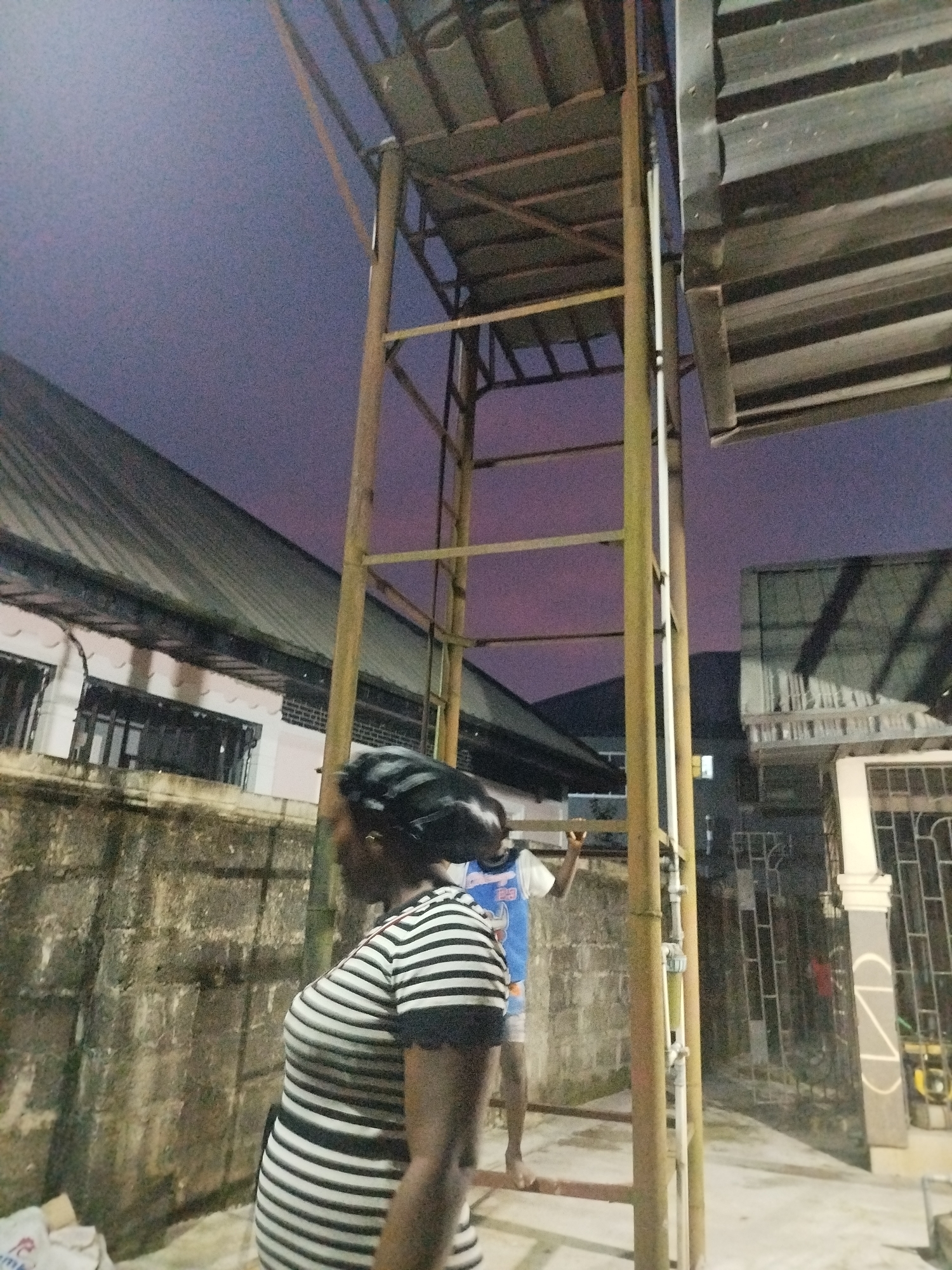In Sapele, Nigeria, an outdoor scene at dusk features a tall, rusty metal ladder structure. A woman wearing a black and white striped shirt stands in the foreground, her back partially to the camera. To her right, a person wearing a blue and white shirt and shorts is positioned on the ladder. The background includes a textured wall, a building with a corrugated metal roof, and other structures typical of an urban Nigerian environment. The sky displays a gradient of purple and pink hues, indicating twilight.