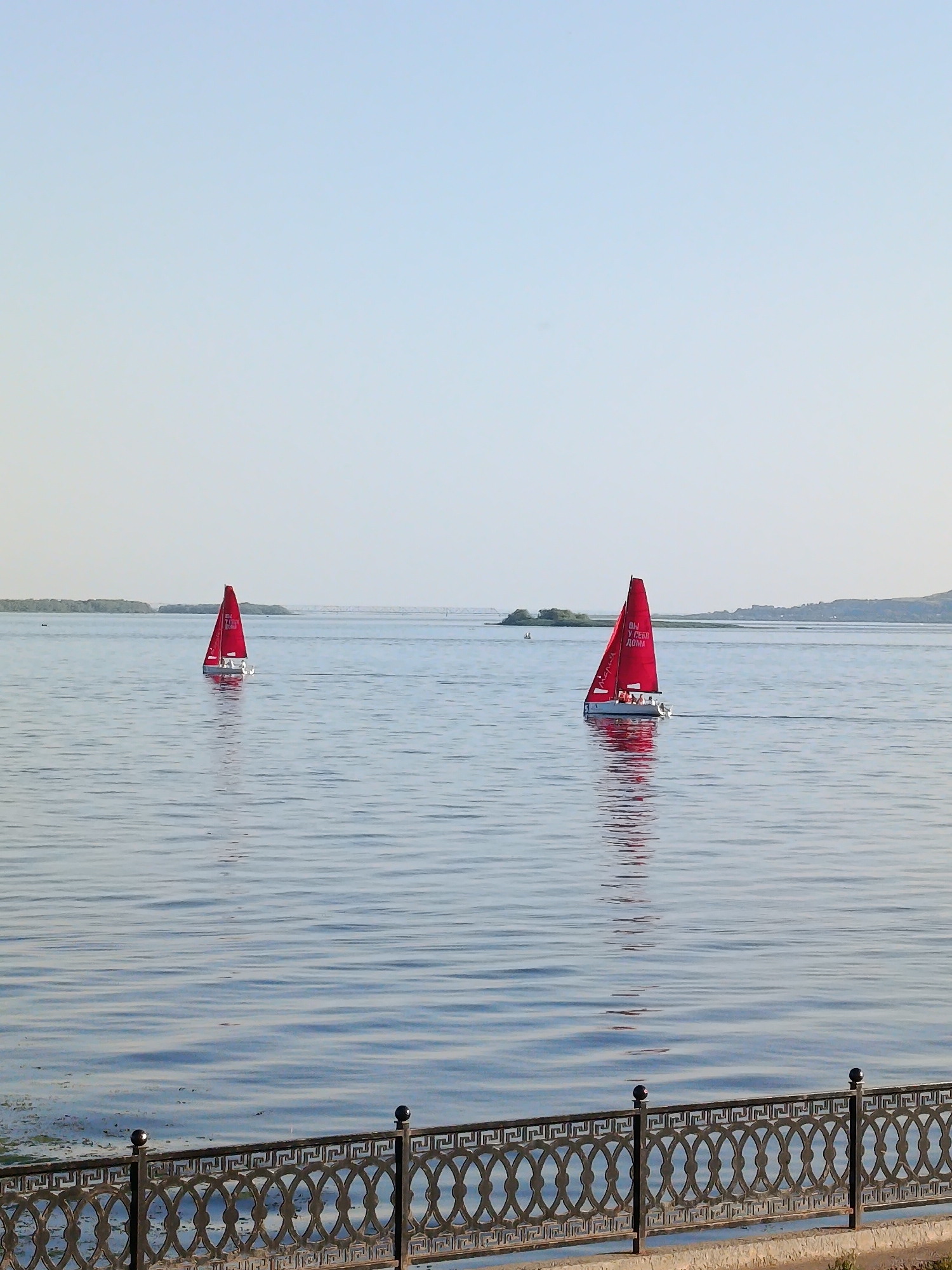 The image captures a serene daytime scene on a wide river, likely the Volga River in Saratov, Russia, given the context. Two sailboats with vibrant red sails are the primary subjects, gliding across the calm, blue water. The sailboat on the right is closer to the viewer and appears to have several people on board. The sails of this boat have some visible white text, which appears to be Cyrillic lettering. The second sailboat is further in the distance, also with a red sail, and appears smaller.
In the background, a low, tree-covered island or landmass is visible on the left, and a longer stretch of coastline with some structures and a bridge can be seen on the right. The sky is a clear, pale blue, suggesting fair weather, and the water reflects the light of the sun, creating gentle ripples. In the foreground, a decorative black metal fence with an intricate geometric pattern runs horizontally across the bottom of the image, separating the viewer's vantage point from the water's edge. The overall mood of the image is peaceful and tranquil, conveying a sense of leisure and the enjoyment of a pleasant day outdoors. The time of day is clearly daytime, likely late morning or afternoon, given the bright, even lighting and the absence of long shadows.