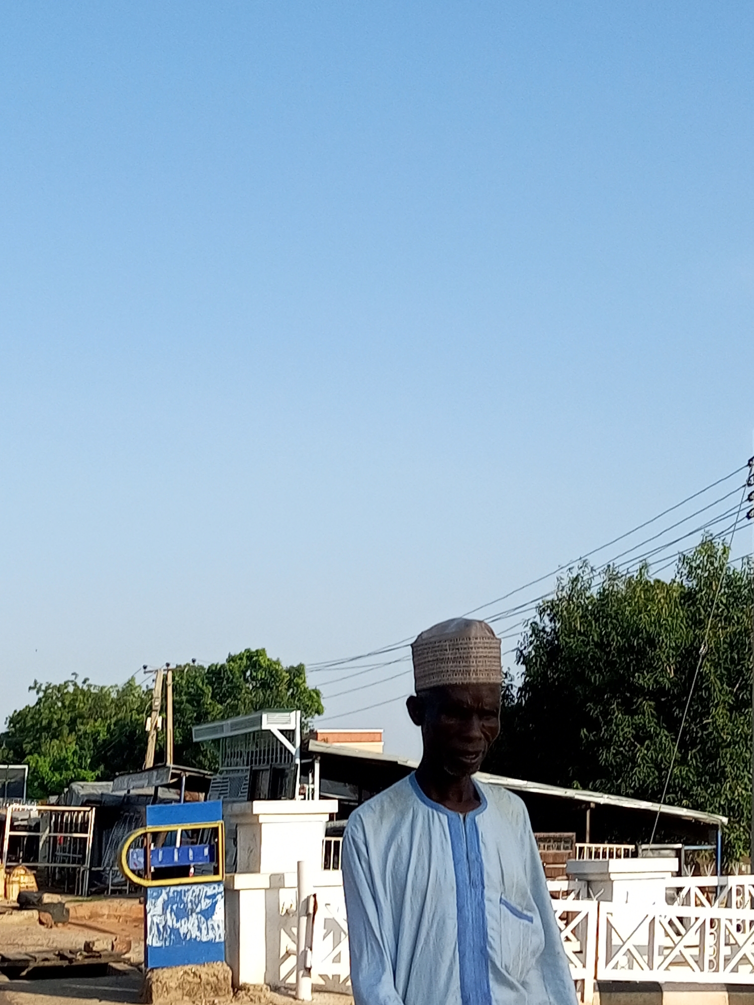 An elderly Black man wearing a light blue traditional African garment and a woven cap stands outdoors, looking to the right. He is positioned in the foreground, slightly off-center to the right. Behind him, to the left, is a blue and yellow sign with some writing on it. Further in the background, there are buildings, trees, and power lines against a clear blue sky. The scene appears to be in an outdoor market or commercial area, likely in Garko, Nigeria, given the context. The overall impression is of a sunny day.