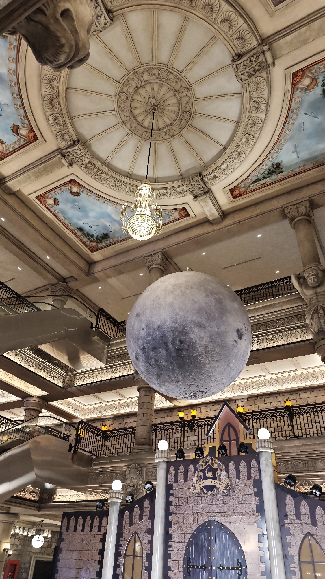 This image depicts a grand, multi-story indoor space, likely a luxury shopping mall, hotel lobby, or entertainment venue, characterized by opulent classical architecture. A massive, spherical replica of the moon hangs prominently in the center of the frame, dominating the mid-ground.
Above, an ornate domed ceiling features intricate relief patterns and a sparkling chandelier suspended from its center. Surrounding the dome are rectangular sections with painted frescoes depicting blue skies and flying birds. The walls are adorned with detailed friezes, columns, and decorative moldings. On the upper left, an escalator with a metallic finish ascends to an upper level, bordered by a black wrought-iron balustrade. On the right, a large column features a sculpted figure, possibly an Atlas or caryatid, supporting the structure above.
In the lower portion of the image, beneath the moon, a detailed castle-like facade is visible. This structure is built from what appears to be stone blocks, featuring arched windows and a large, dark-colored entrance gate with metal studs. Above the gate, a crest displays the text "MOON KINGDOM" and "Est. 2017". Spotlights and decorative globe lights illuminate the castle display. No people are visible in the scene, suggesting a focus on the architectural grandeur and the themed exhibition. The overall impression is one of a fantastical and immersive environment, with the moon as a central, awe-inspiring element.