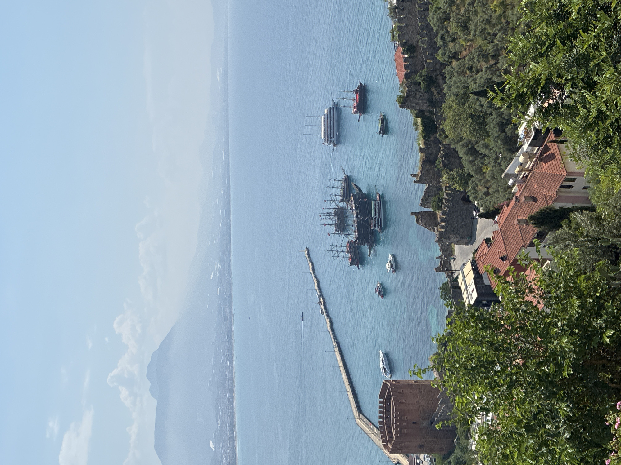 This is a high-angle, eye-level shot of the Red Tower (Kızıl Kule) and the harbor of Alanya, Turkey, on a sunny day. The Red Tower, a prominent historical landmark, is a circular, reddish-brown stone structure located on the left side of the image. Adjacent to the tower, a long, concrete pier extends into the bright blue sea. Several boats are docked or anchored in the harbor, including a large white yacht, smaller motorboats, and several traditional wooden sailing vessels, some of which appear to be pirate-themed tourist boats.
The sea is calm with clear, turquoise waters near the shore and a deeper blue further out. In the background, the city of Alanya stretches along the coastline, with buildings and houses visible. Beyond the city, distant mountains are shrouded in a light haze, with a few wispy clouds in the pale blue sky.
The foreground of the image is filled with lush green trees and vegetation, with the red-tiled roofs of buildings peeking through. Some sections of ancient stone walls, part of the Alanya Castle complex, are visible on the right side of the image, integrating with the natural landscape. The overall scene depicts a picturesque view of the historic harbor and coastal landscape of Alanya.