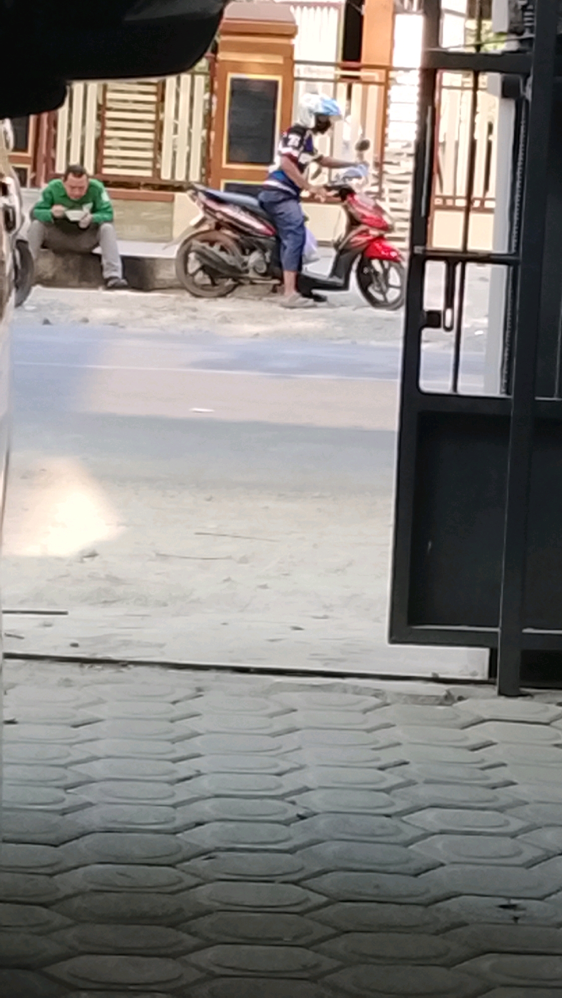 In Slawi, Indonesia, a man wearing a green long-sleeved shirt and grey pants sits on a concrete curb, eating from a white bowl. Next to him, a person wearing a blue and white striped shirt, blue pants, and a white helmet is seated on a red motorcycle. The motorcycle is parked on a dirt or gravel surface. Behind the individuals, a building with light-colored walls and a patterned decorative facade is visible. The foreground of the image shows a patterned concrete walkway. The overall scene appears to be an outdoor area in a residential or commercial setting within the city.