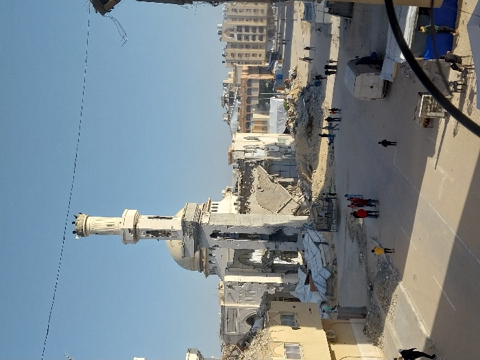 The image depicts a scene of extensive destruction in Khan Yunis, Palestinian Territory, under a clear blue sky. A large mosque, identified by its prominent minaret and dome, stands centrally in a state of severe damage; its dome is partially collapsed, and significant portions of its walls and roof are missing, exposing internal structures and large piles of debris at its base. The minaret, though still erect, shows visible damage and a small group of individuals is visible near its upper section.
The foreground and midground reveal a street and surrounding areas covered in a mix of asphalt, sand, and large quantities of concrete rubble, twisted metal, and other building debris. To the left, a partially damaged multi-story building with a light-colored facade is visible.
Numerous people are dispersed across the scene: a group of approximately seven individuals, some wearing red jackets, stands on the paved section of the street in the mid-left. Other individuals are seen walking across the debris-strewn ground and paved areas. A person in a yellow shirt walks towards the left foreground, and another in black is on the far left. In the lower right foreground, several people are seated on plastic chairs beneath a blue tarp, beside a small table. A white panel van is parked on an unpaved section of ground. Beyond the primary mosque structure, additional multi-story buildings are visible in the background, some appearing intact and others potentially damaged. Thin black wires or cables stretch across the upper portion of the sky. The perspective is from an elevated vantage point looking down onto the street level.