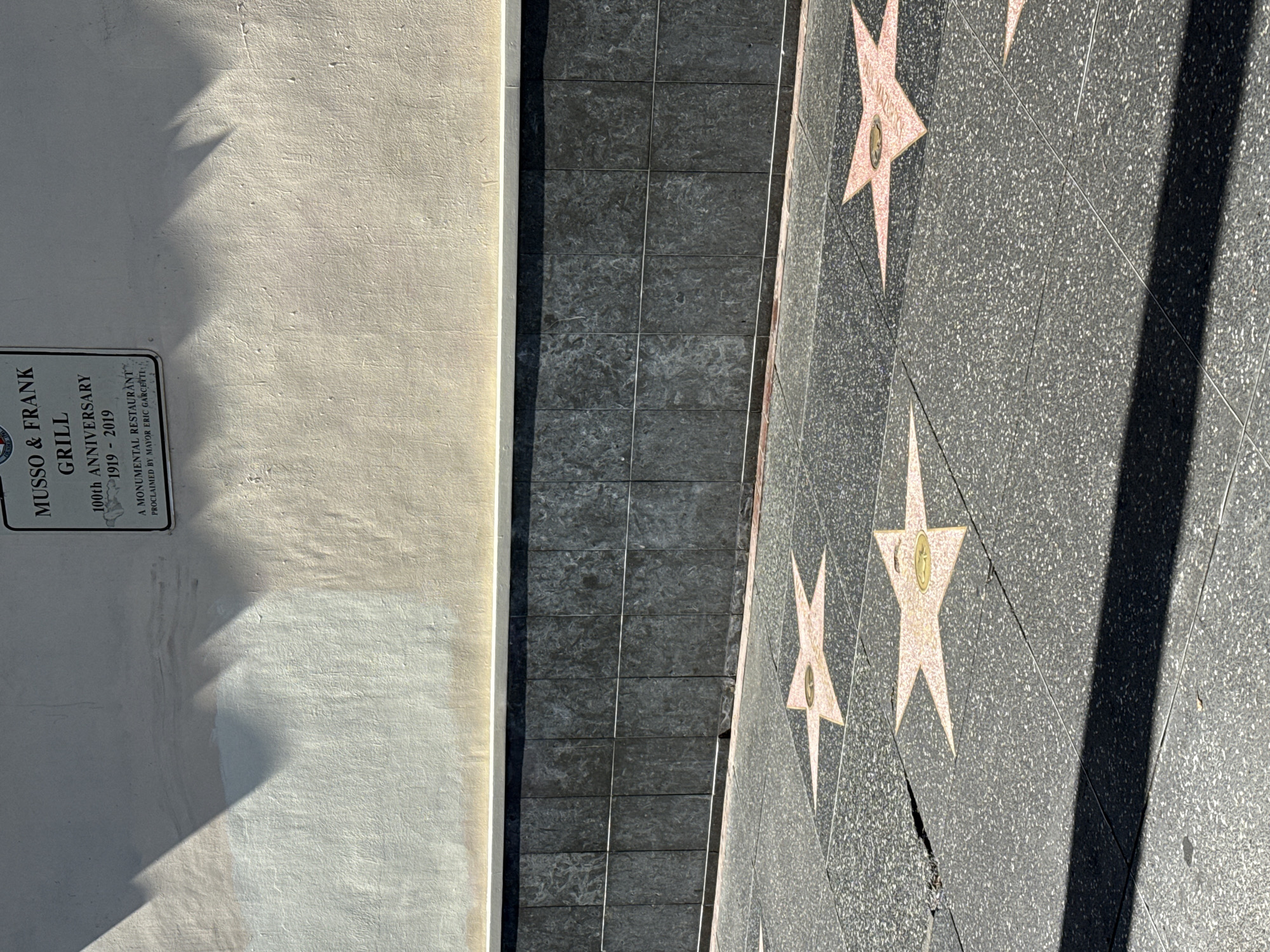 The image shows a portion of the Hollywood Walk of Fame in Los Angeles, California. In the foreground, several stars are embedded in the sidewalk, each representing an honoree. One prominent star is pink and features a bronze emblem and text, though the name is not clearly legible. The sidewalk itself is made of dark gray speckled material with visible grout lines. To the right, another pink star is partially visible. A dark shadow, likely cast by a building or tree, cuts across the lower portion of the image. Behind the sidewalk, there is a dark, gray, marble-like tiled wall. Above this wall, a light-colored stucco wall is visible, with areas of discoloration and the faint imprint of shadows, possibly from palm fronds or architectural elements. A black and white sign is mounted on the stucco wall, displaying text that reads "MUSSO & FRANK GRILL" and "100th ANNIVERSARY 1919 - 2019". Below this, further text on the sign states "A MONUMENTAL RESTAURANT" and "PROCLAIMED BY MAYOR ERIC GARCETTI". The scene is illuminated by bright sunlight, creating distinct shadows.