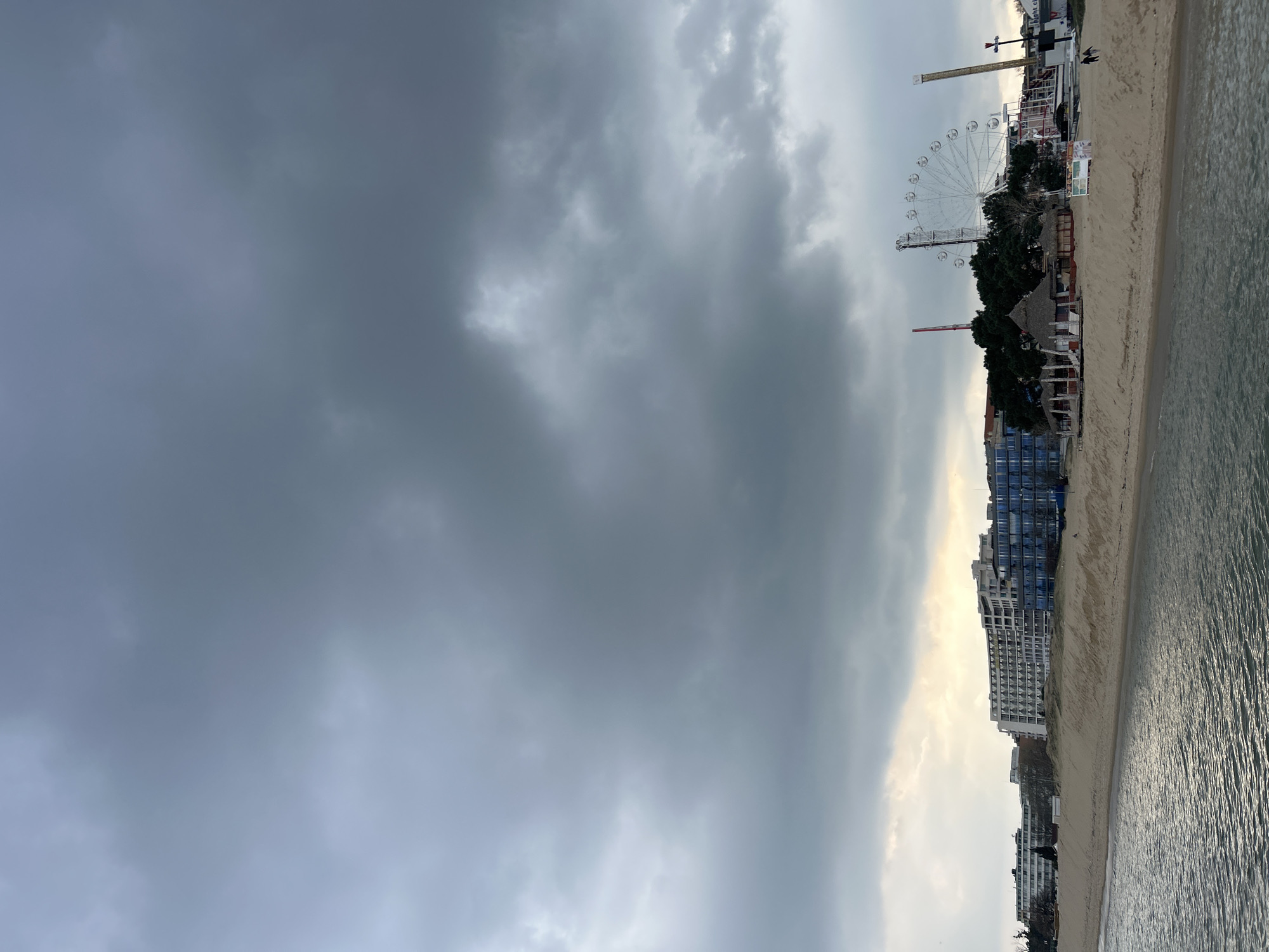 The image depicts a beach scene under a cloudy sky. A tall building and a ferris wheel are visible along the shoreline, adjacent to an amusement park. The location is likely a coastal area in a city; the specific name of city is Unknown.