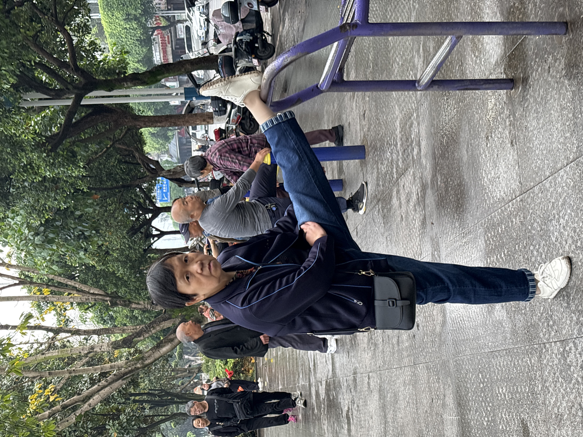 The image depicts a woman in the foreground stretching her leg on a purple metal bar in what appears to be a public park or recreation area. She is wearing a navy blue jacket, jeans with the cuffs rolled up to reveal striped lining, and white sneakers. A small black bag hangs from her shoulder. The woman looks directly at the camera.
Behind her, several other people are also stretching on similar bars. One man is wearing a grey shirt and another a plaid shirt. Further back, other people are walking along a path.
The ground is paved and wet, suggesting it may have recently rained. Trees are visible on the left side of the frame, with vehicles, including motorcycles, parked along the street in the background.
The location is Jiangjin, China.