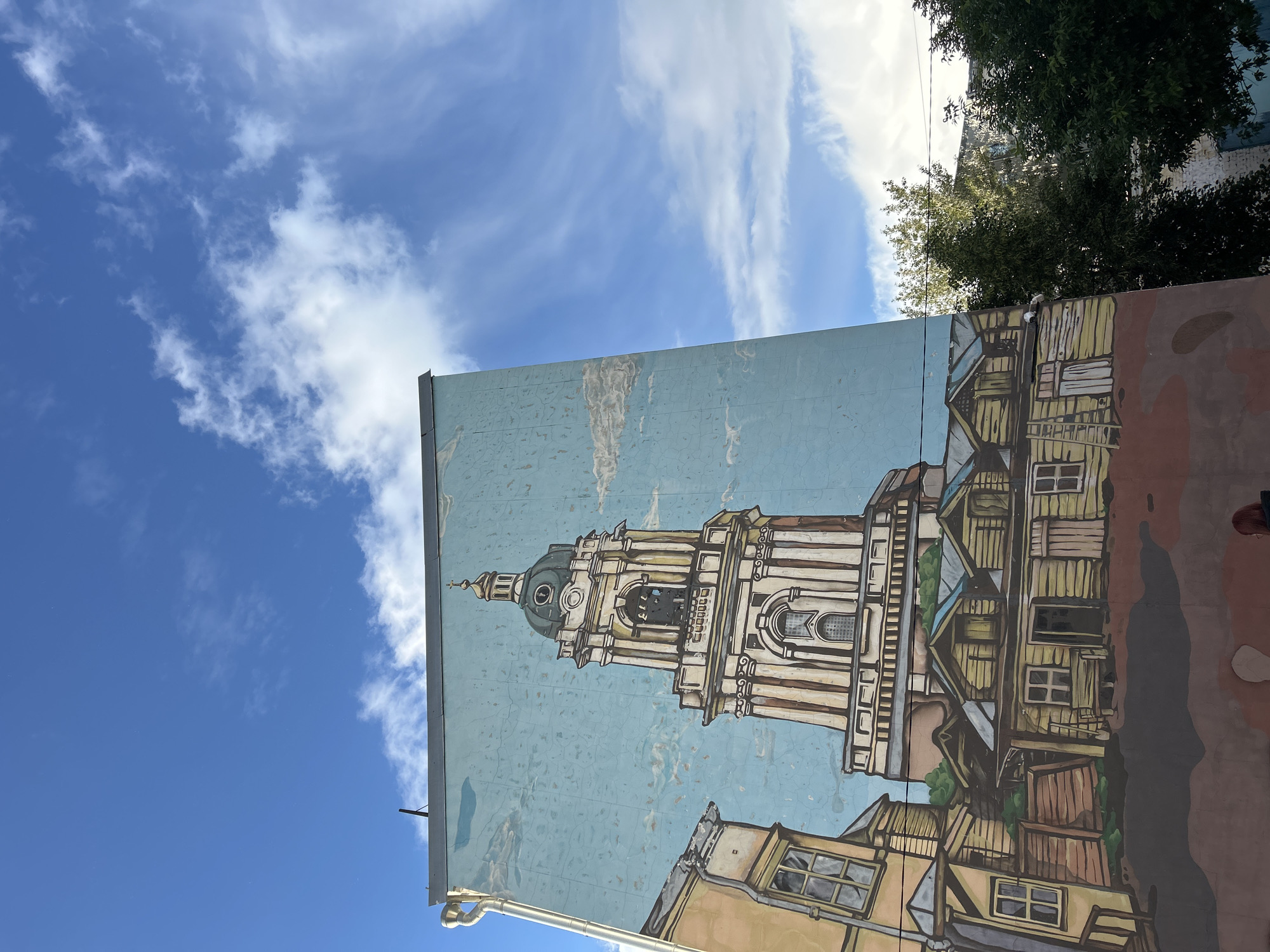 A mural depicting a church tower and surrounding buildings is painted on the side of a building. The sky with white clouds occupies the upper left portion of the image. The mural extends from the bottom of the frame upwards, showcasing architectural details of the church tower and adjacent structures. The buildings appear to be rendered in a stylized, illustrative manner. Green trees are visible to the upper right. The photo is taken from a low angle, looking up at the mural.