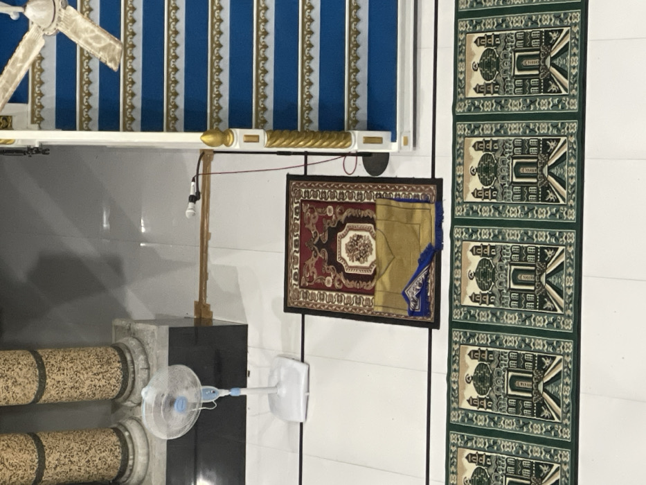 A room features a decorative ceiling with blue and white stripes and gold trim, under which hangs a white ceiling fan. A brown and gold carpet hangs on the wall next to a green prayer rug with repeated geometric patterns. Below, a pedestal supporting a microphone is visible, along with a free-standing fan and decorative pillars with a patterned surface. The wall is white with thin, vertical black lines.